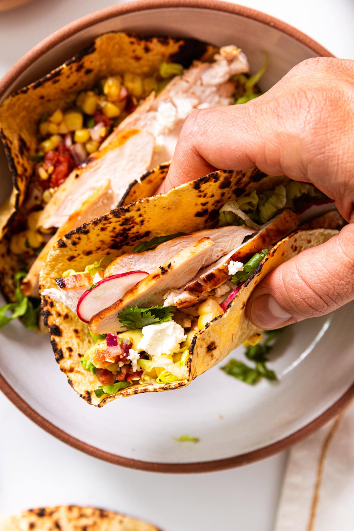 Hand holding a chicken taco with slices of radish on top along with crumbly cheese with another taco in stone bowl below.