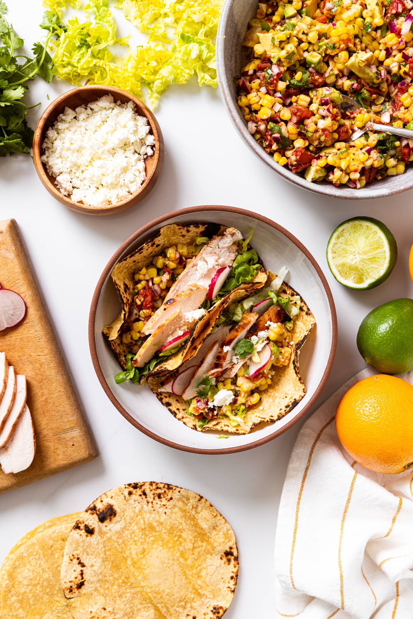 Large stoneware bowl filled with two chicken tacos topped with corn salsa and lettuce with lots of toppings sitting all around on white countertop.