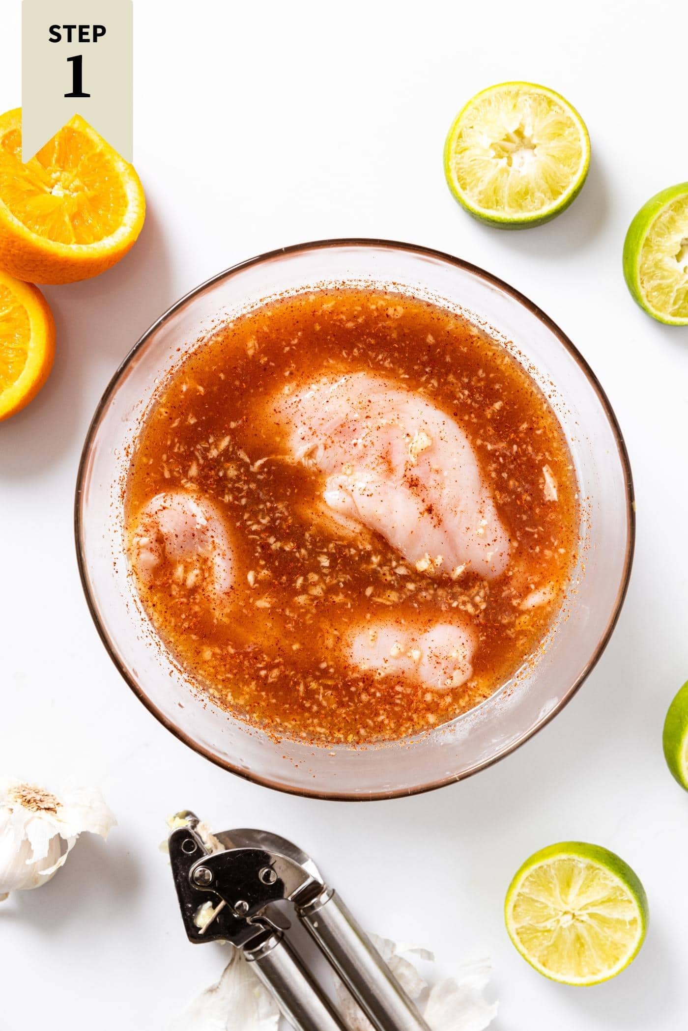 White countertop with glass bowl filled with marinade liquid with raw chicken breasts inside with used limes and oranges all around.