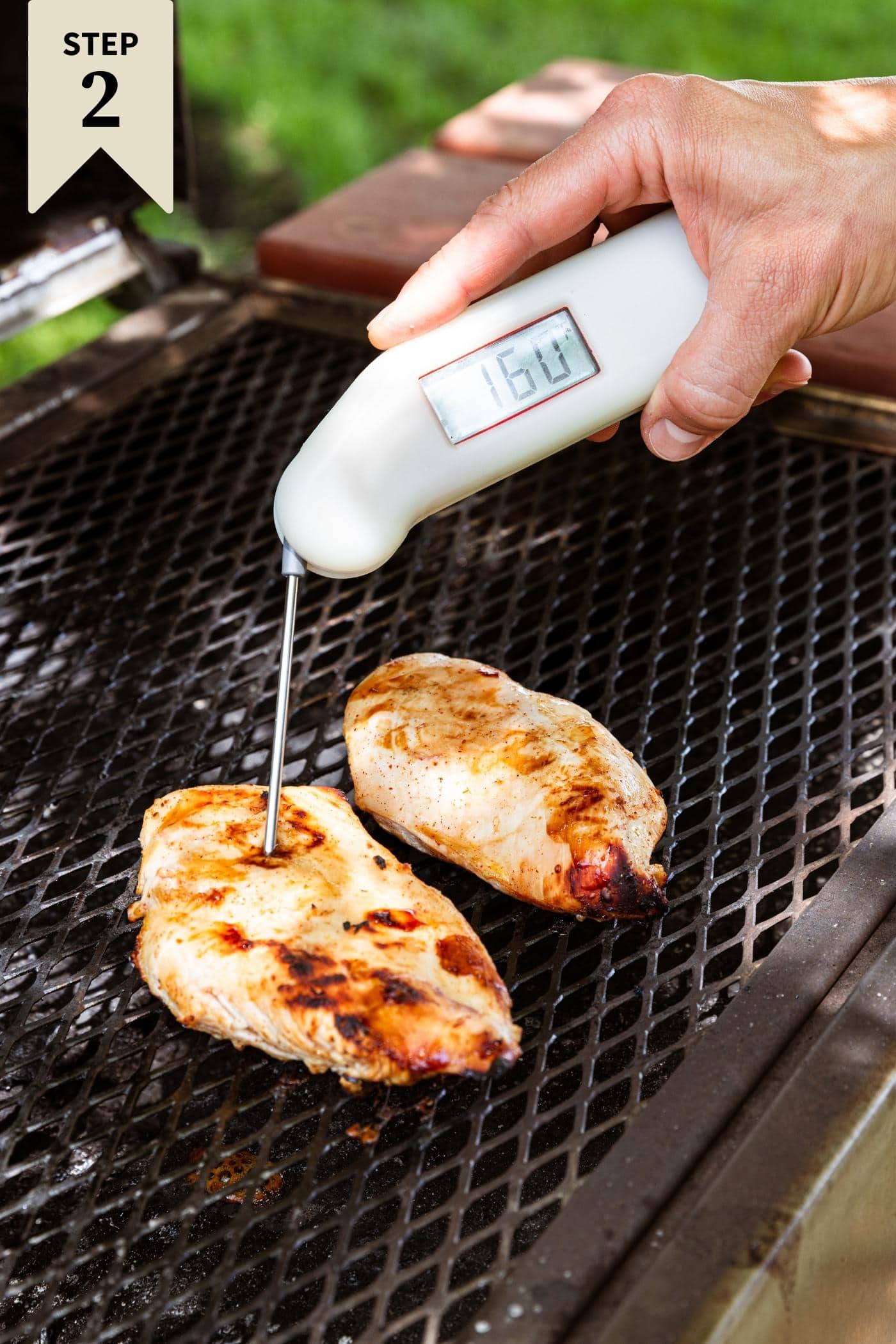 Hand holding instant read thermometer with end in middle of grilled chicken breast charbroiled on an open grill.
