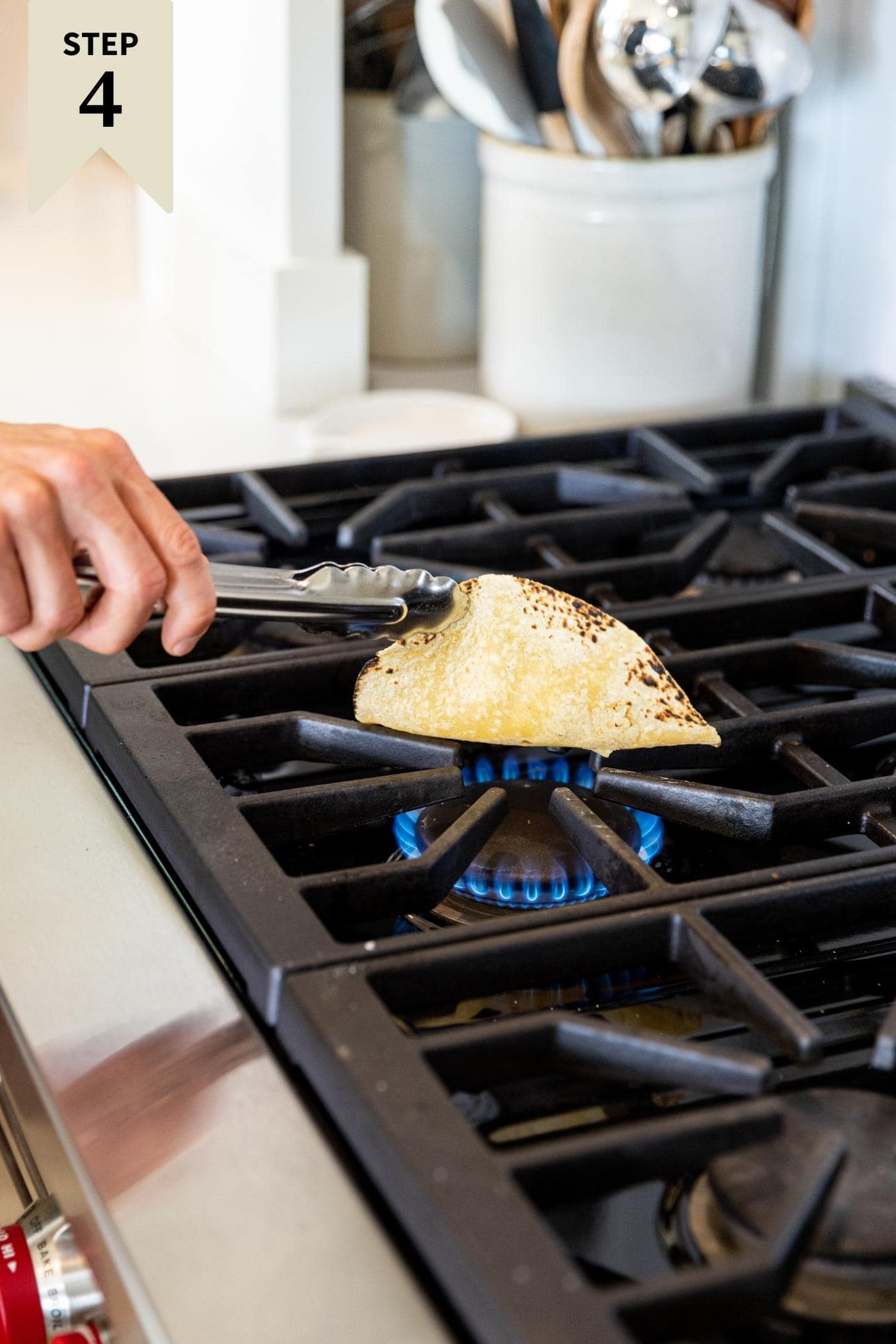 Hand holding tongs holding corn tortilla charring it slightly on open gas grill stove.
