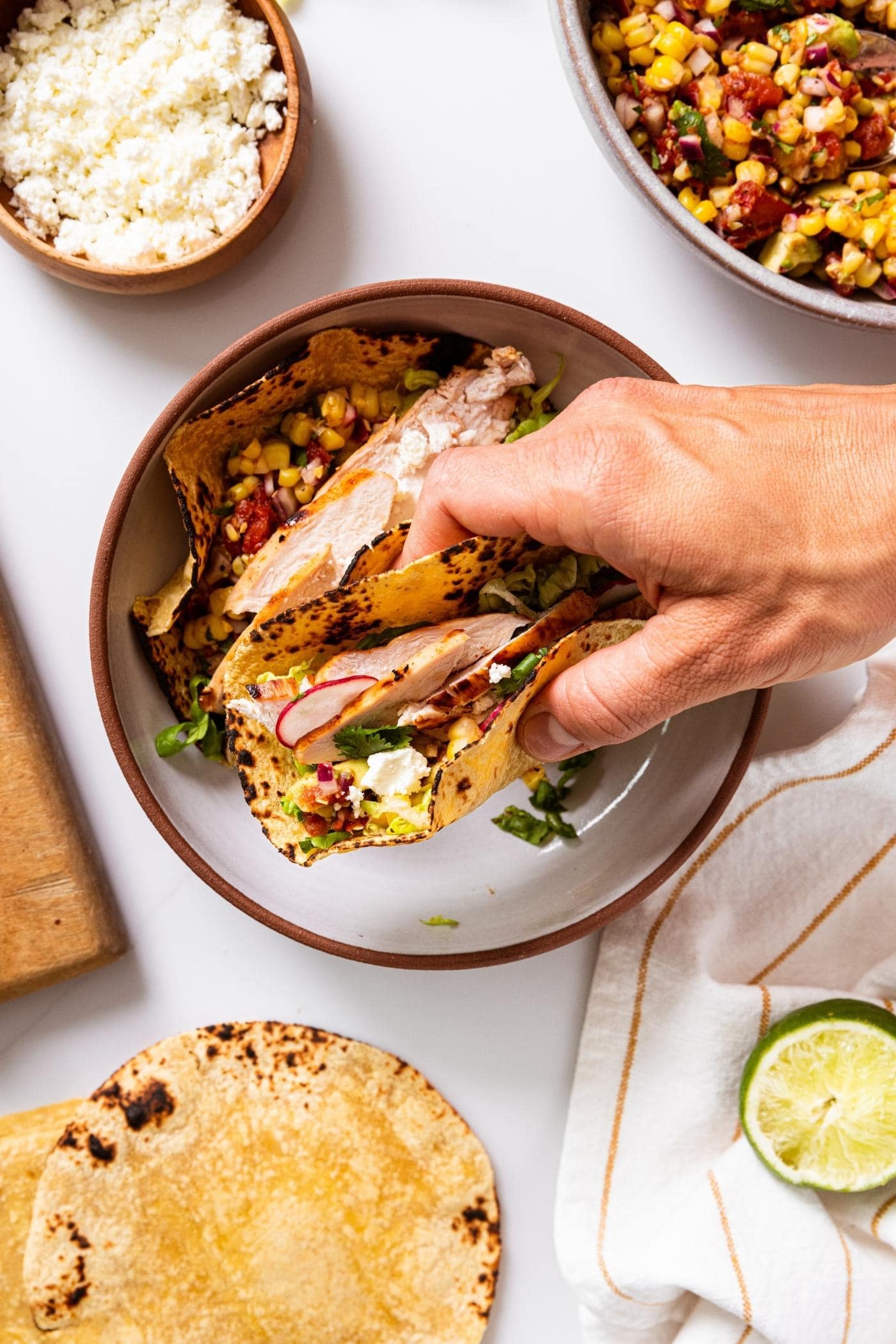 Hand holding folded chicken taco over brown bowl with other bowls around holding cheese and corn salsa.