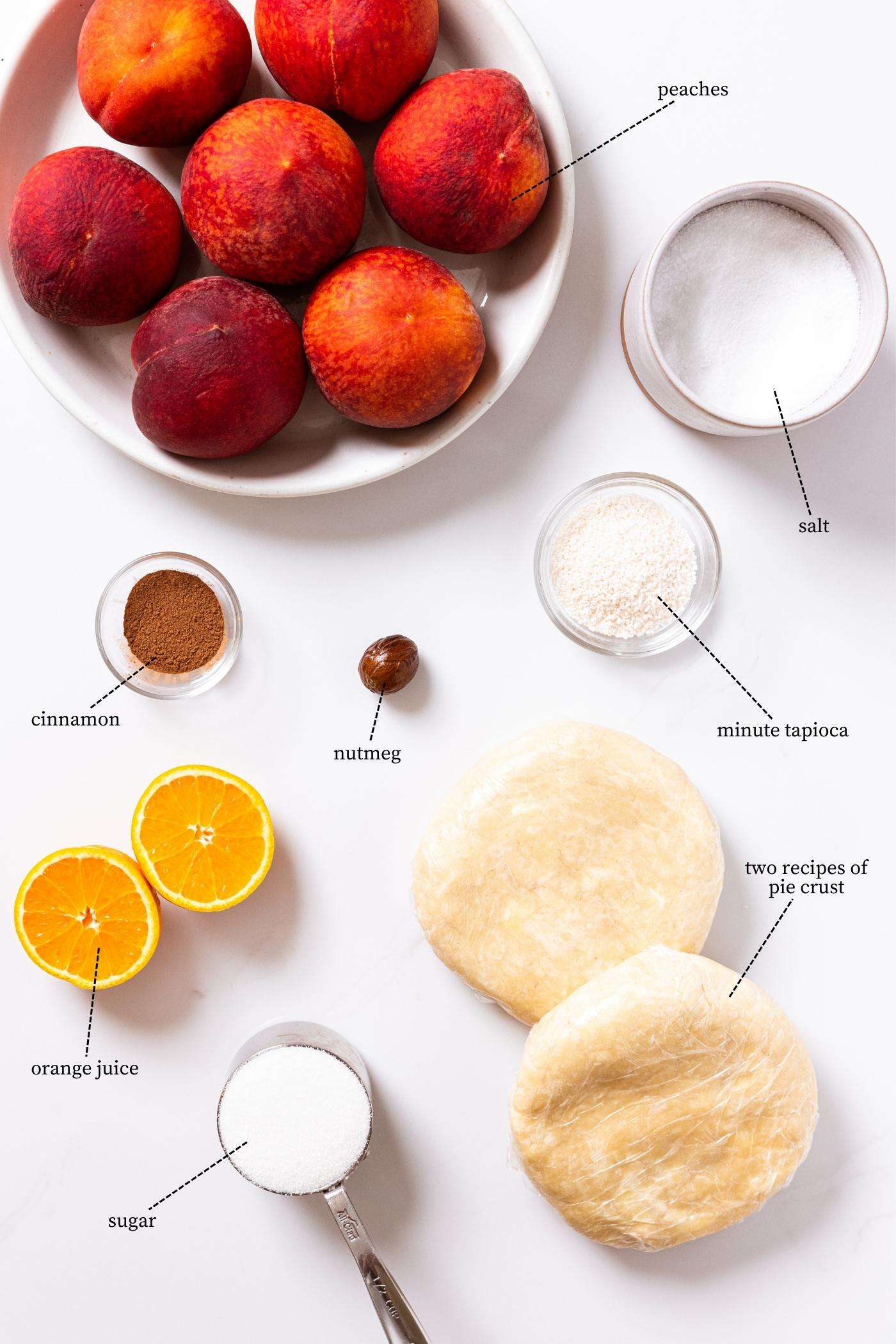 White countertop filled with all ingredients needed to make a peach pie including peaches, pie crust, oranges, cinnamon, nutmeg, and more.