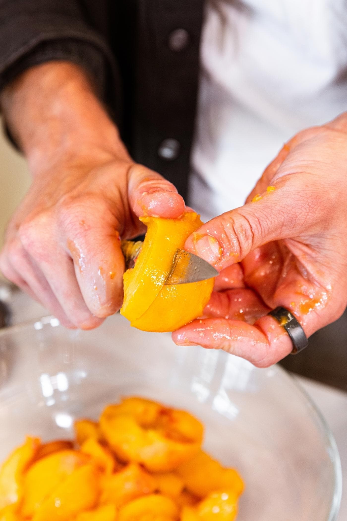 Two hands holding a peach slicing into small slices with bowl of peaches below all on white countertop.