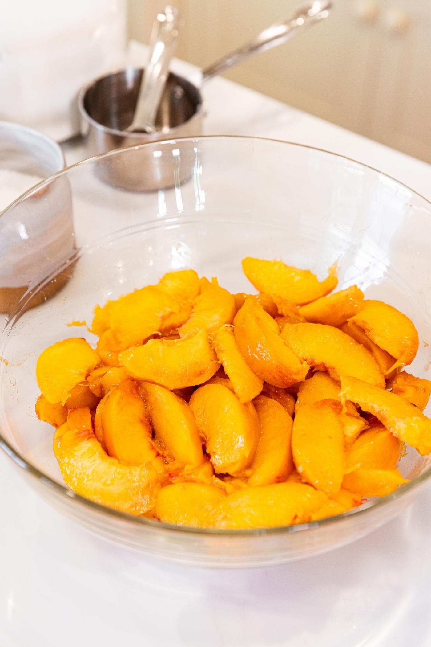 Glass bowl sitting on white countertop filled with peach slices with metal measuring cups in background.