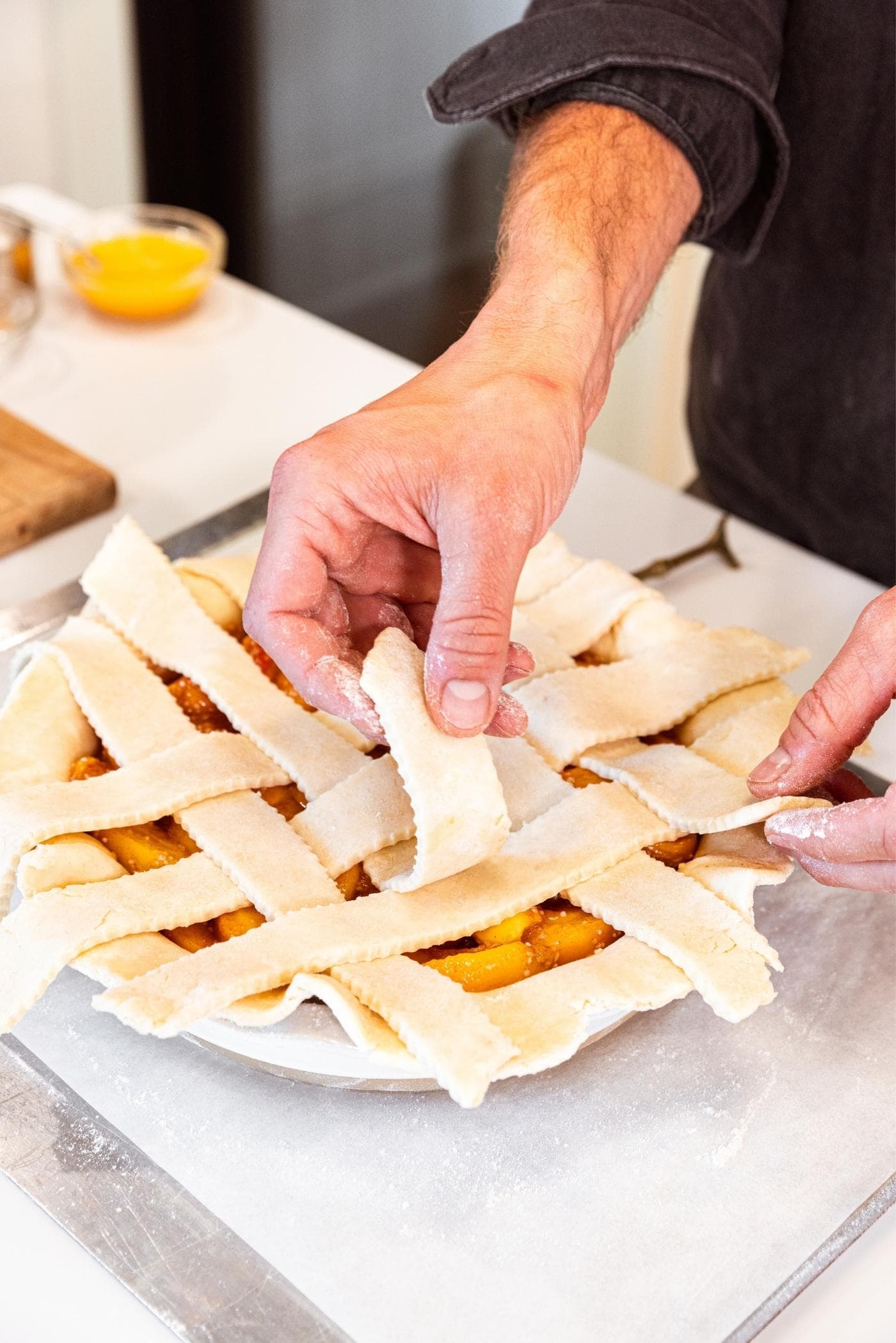 Hand laying lattice on top of peach pie sitting on white countertop.