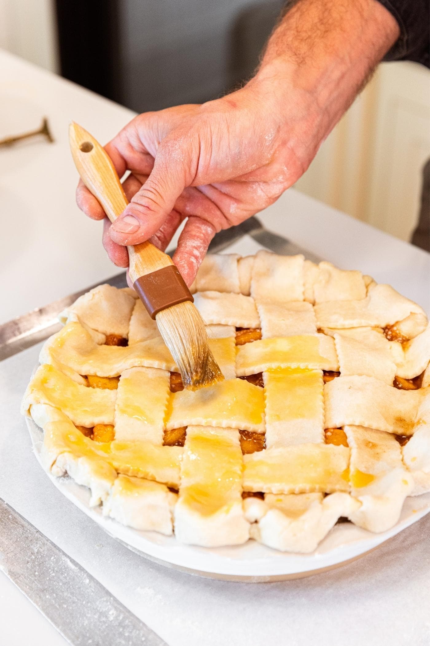 Hand brushing egg wash onto top of peach pie using a brush.