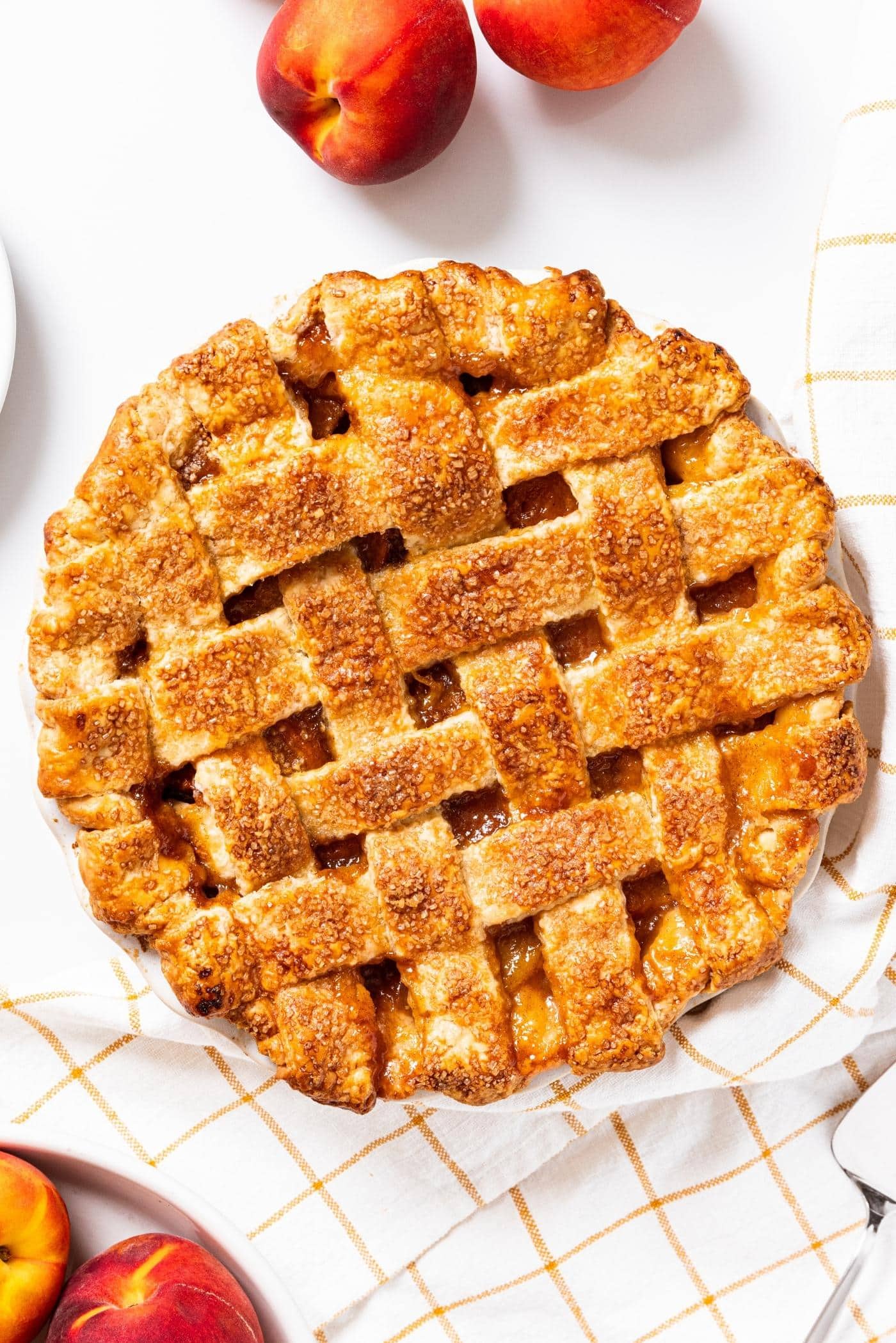 Baked peach pie sitting on white countertop with a few peaches sitting around with checkered yellow dish towel underneath.
