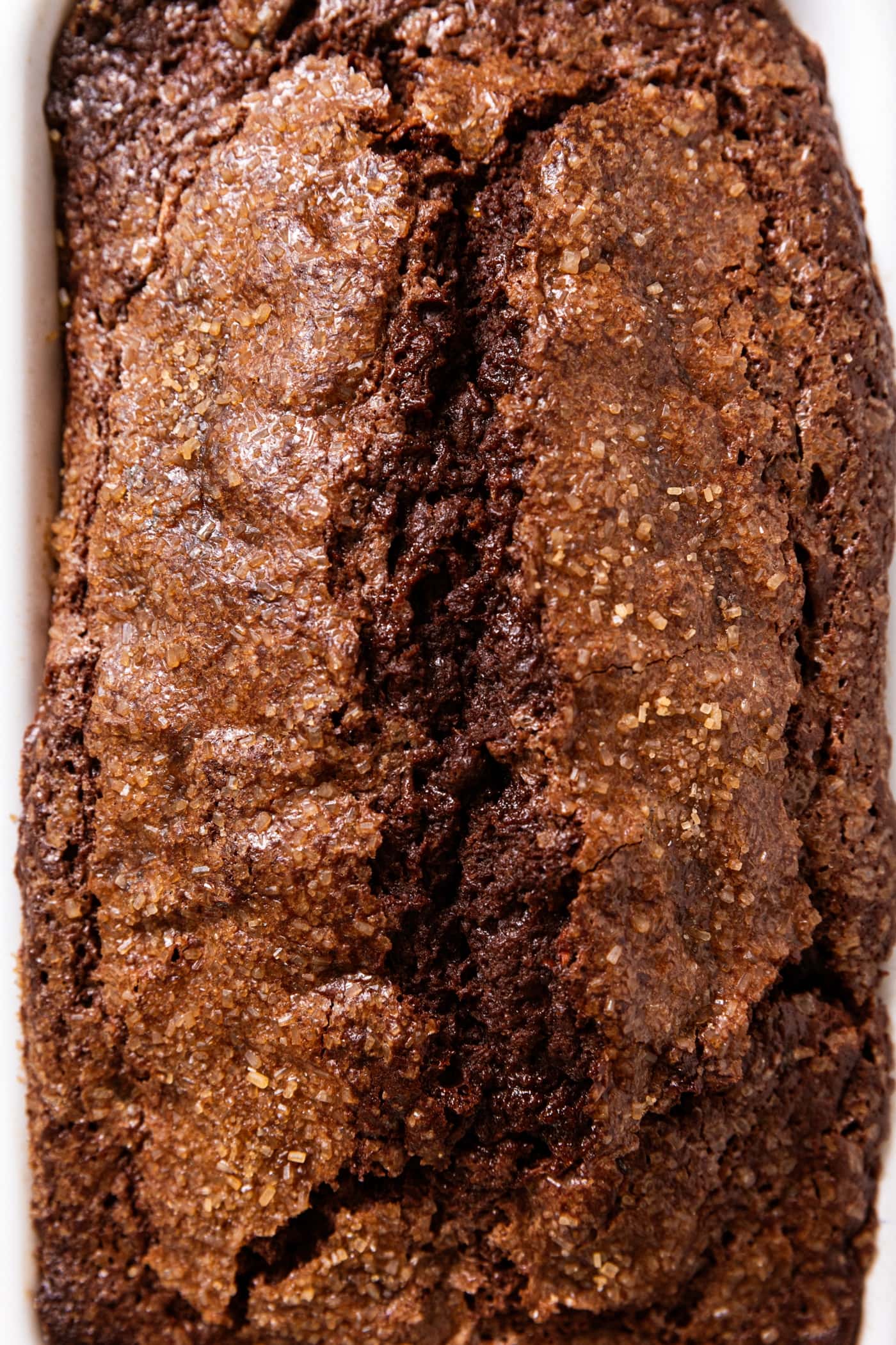 Chocolate zucchini loaf of bread in white baking dish showing the deep crack on top with crackly topping from sugar.