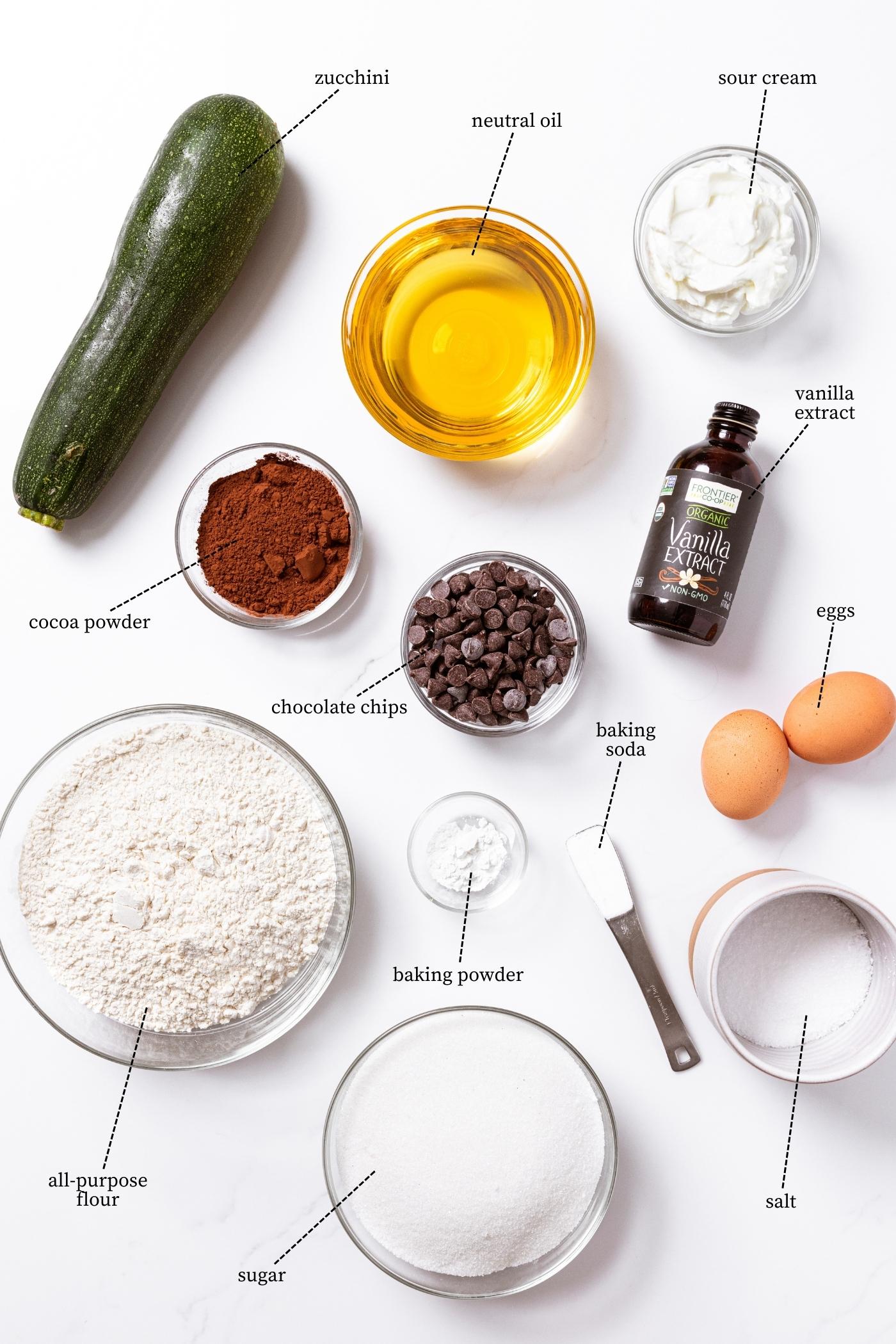 White countertop filled with everything needed to make chocolate zucchini bread including zucchini, flour, sugar, salt, oil, cocoa powder, vanilla, eggs, and more.