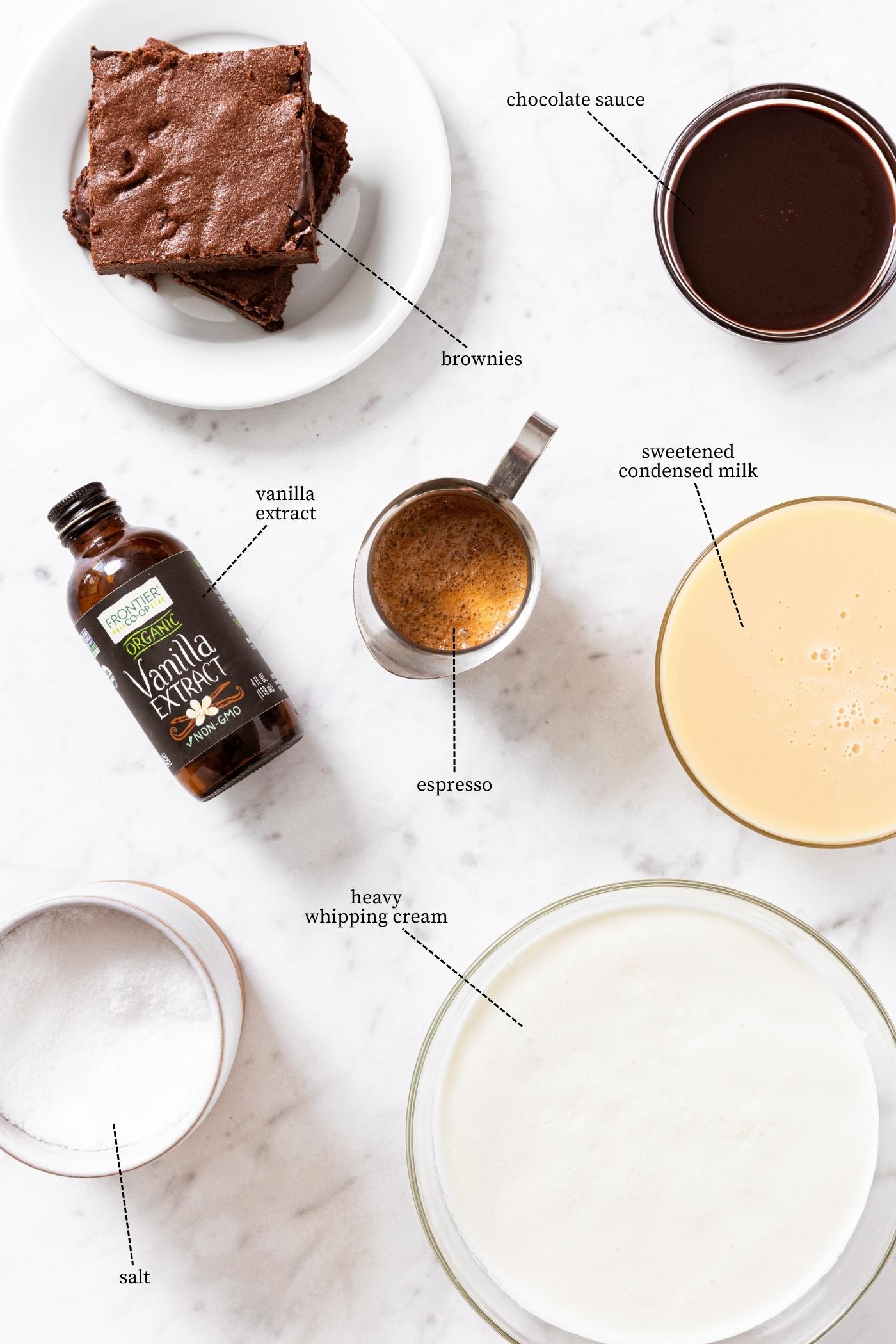 White marble countertop filled with all the ingredients needed to make espresso flavored ice cream including brownies, chocolate sauce, espresso, cream, and more.