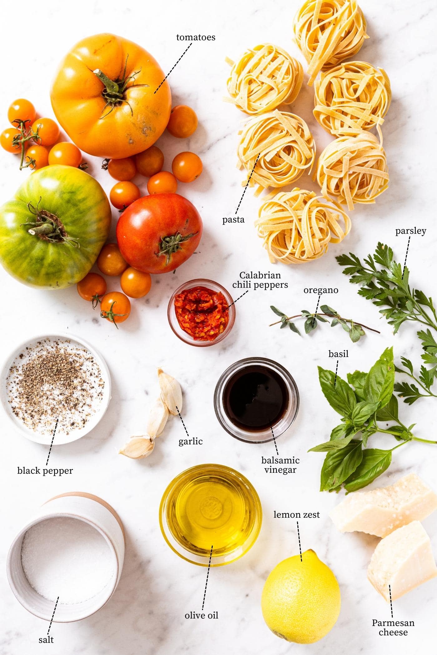 Marble countertop filled with everything needed to make a fresh tomato pasta recipe including tomatoes, pasta, herbs, oil, lemon, cheese, and more.