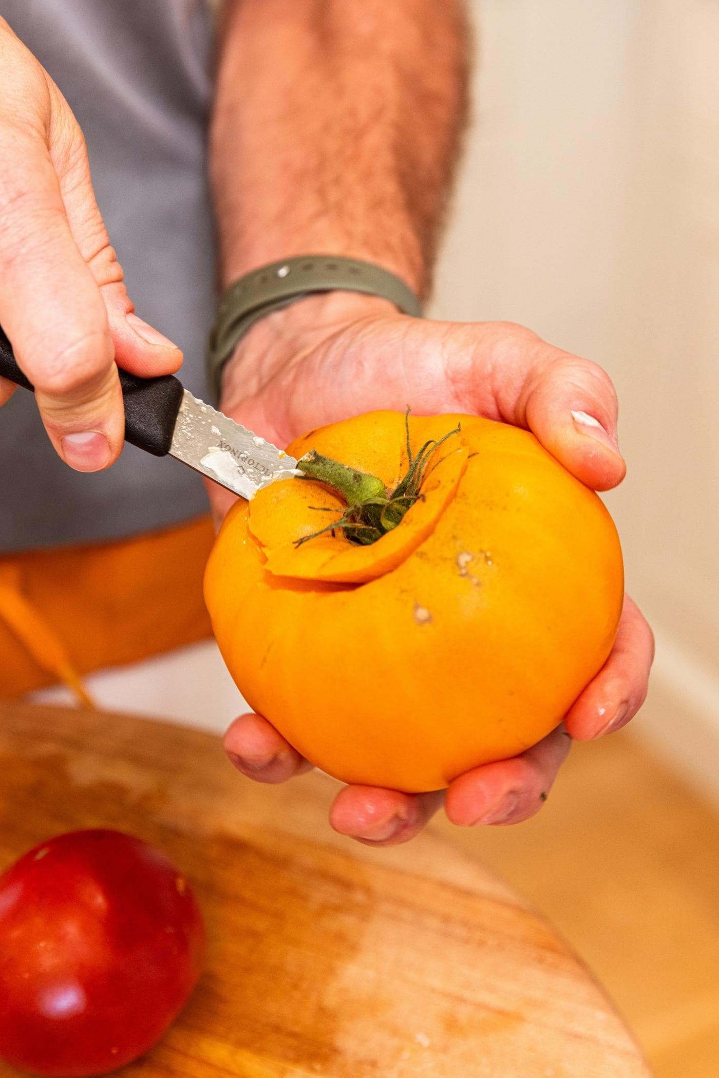 Hand using knife to core the center of a large yellow tomato on top of a cutting board.