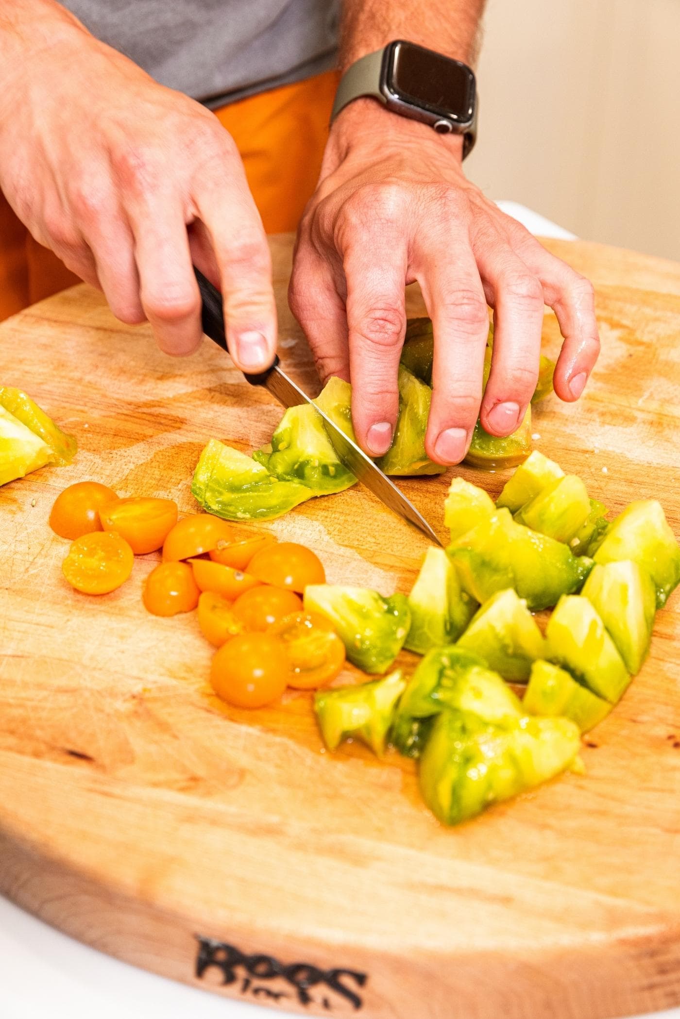 Hand slicing green tomato with a knife on cutting board with yellow cherry tomatoes also on board.