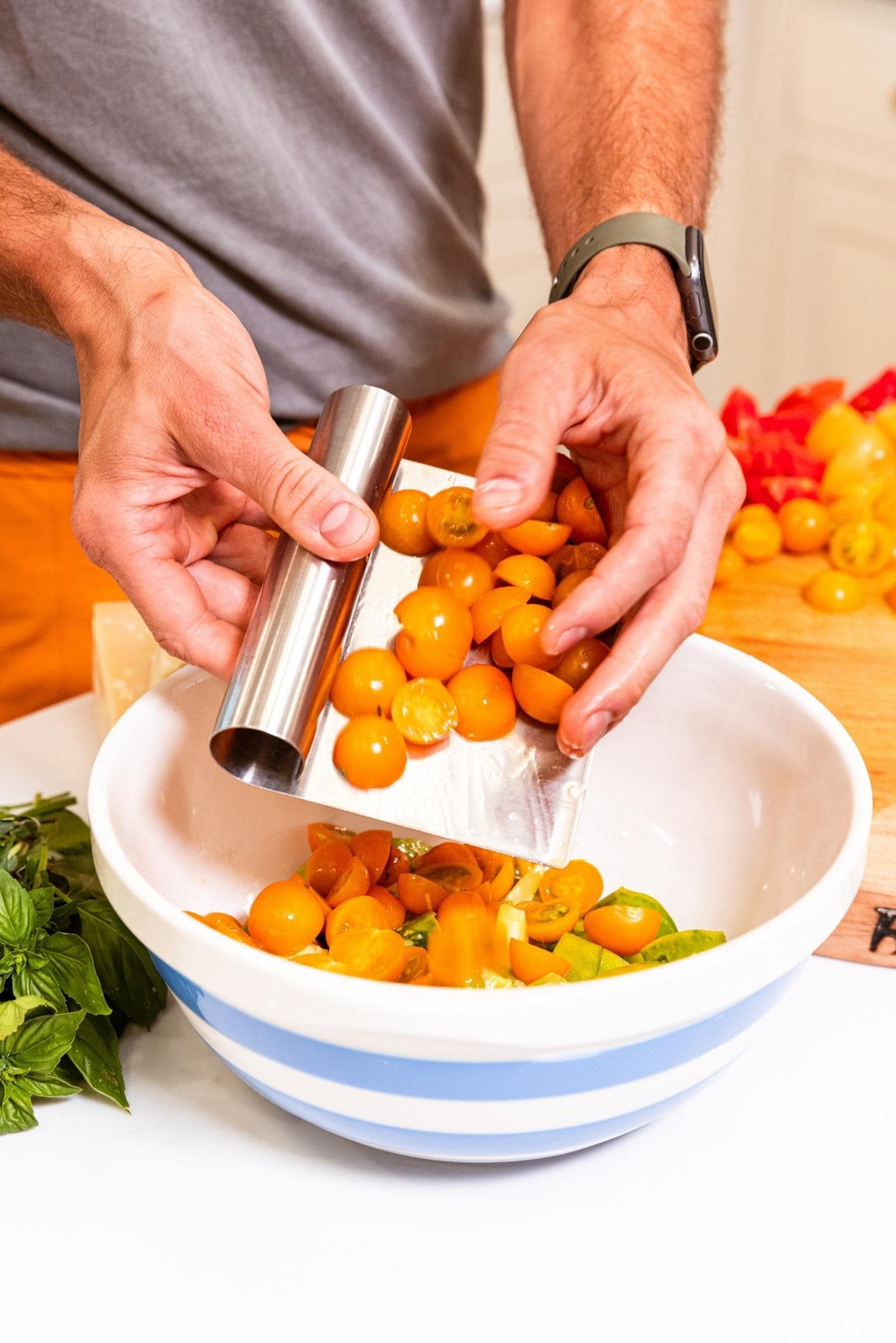 Hands using metal bench scraper to place sliced tomatoes of various colors into large white bowl with blue rings.