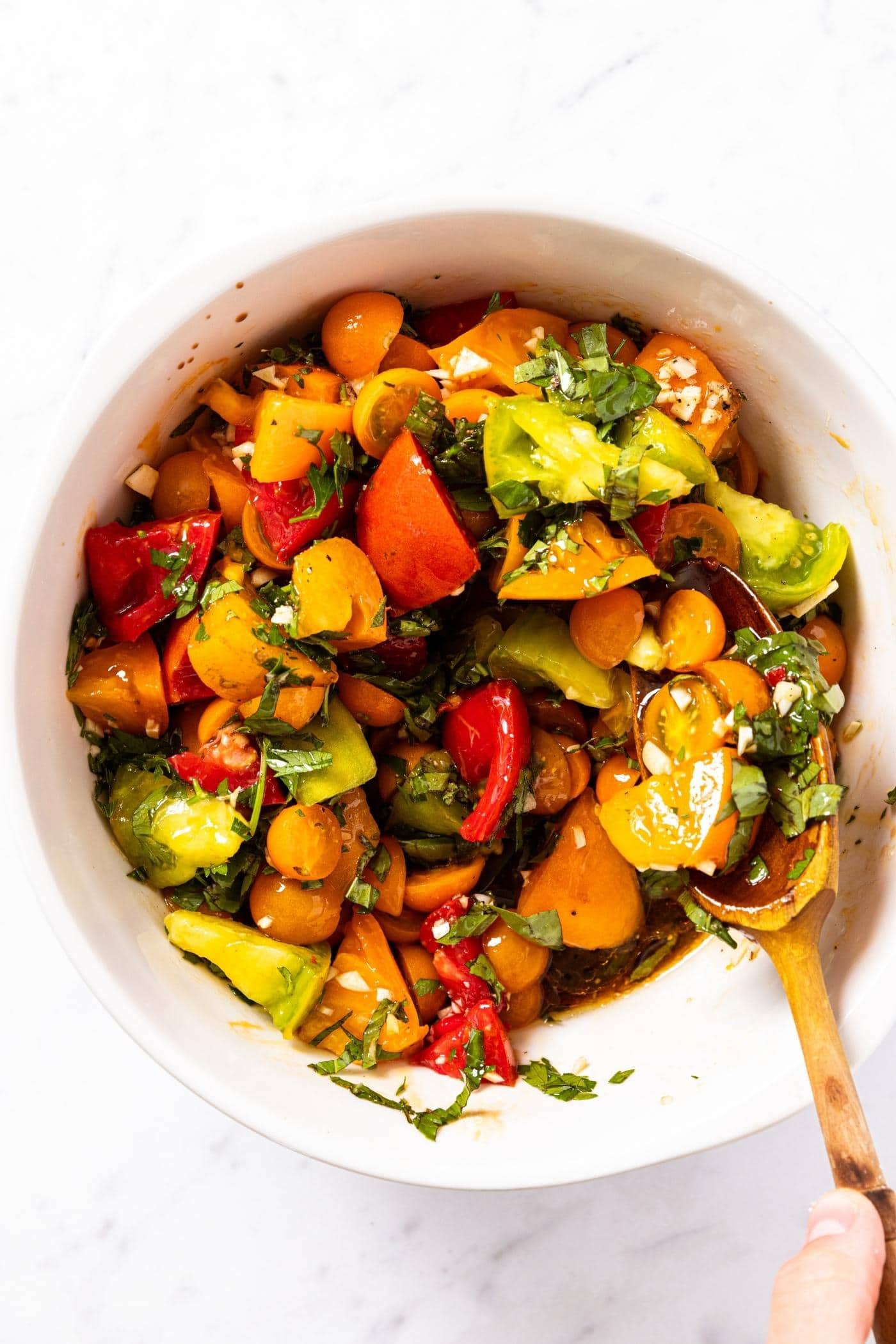 Wooden spoon stirring together tomato chunks with fresh herbs and garlic all on white marble surface.
