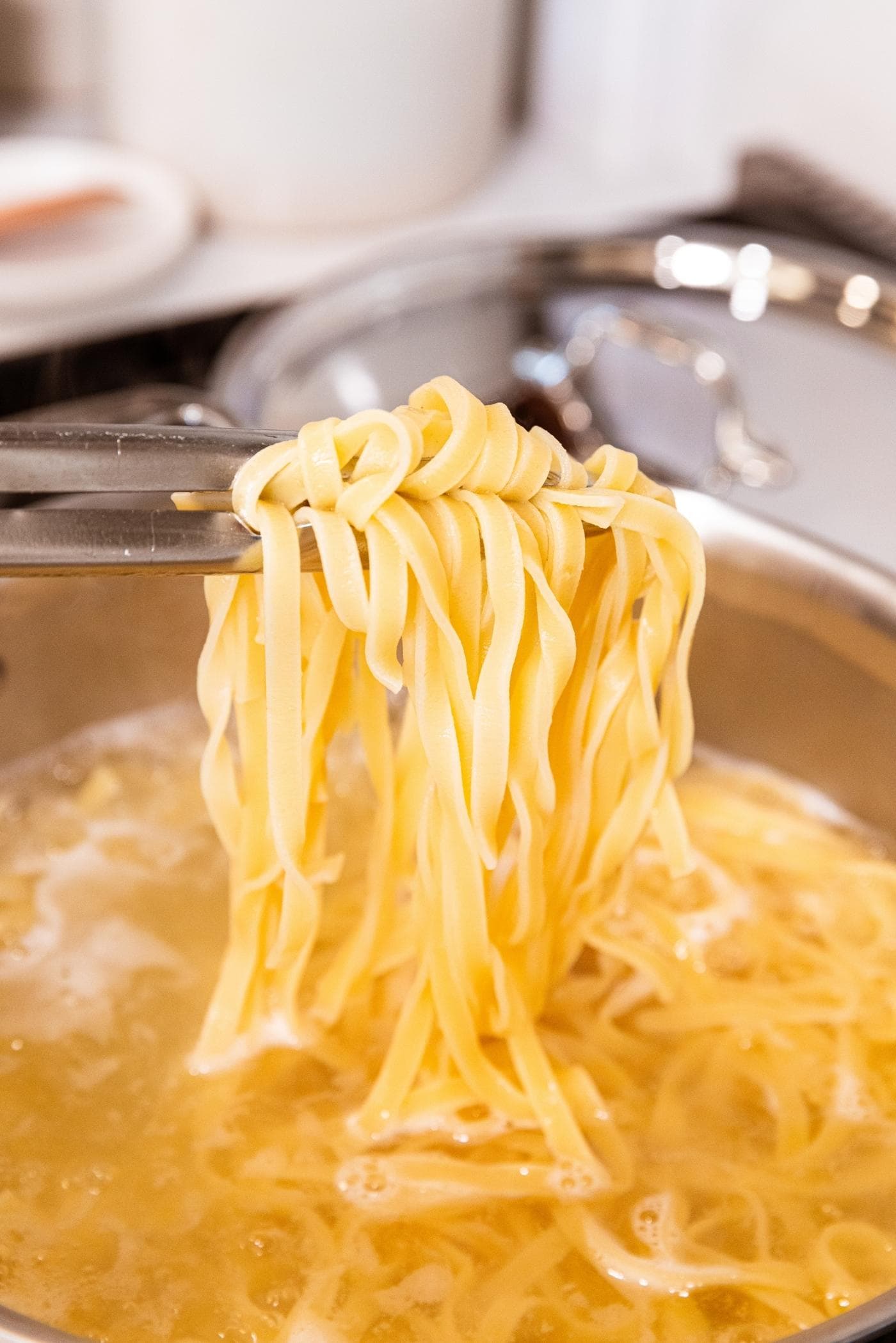 Metal tongs picking up cooked pasta noodles from boiling water in kettle on stovetop.