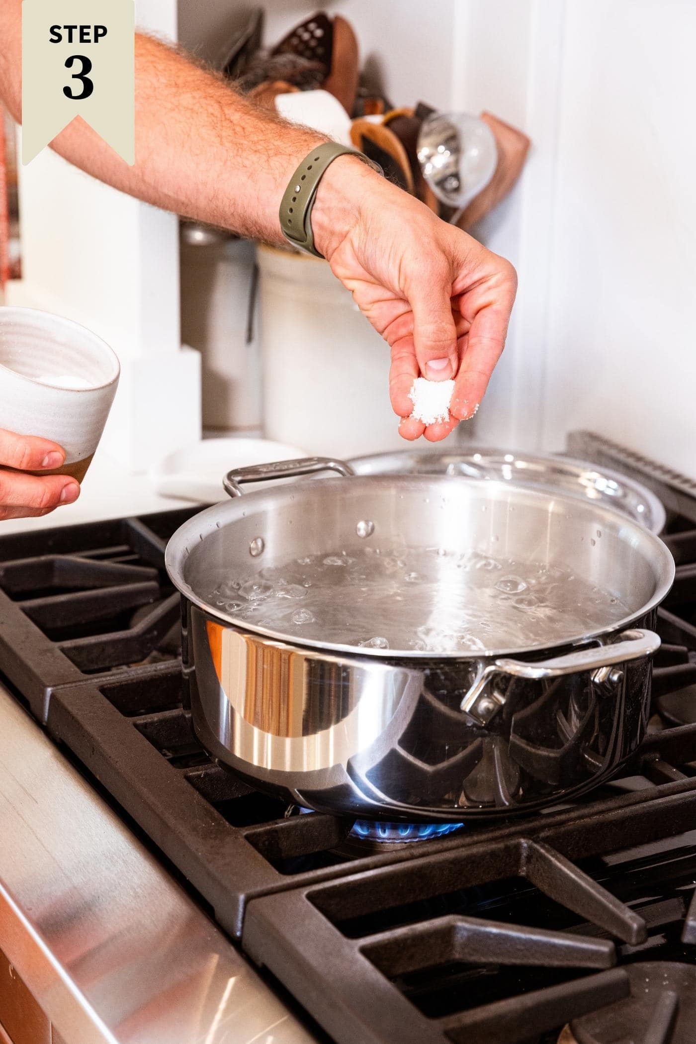 Hand sprinkling salt into large kettle on stovetop filled with water.