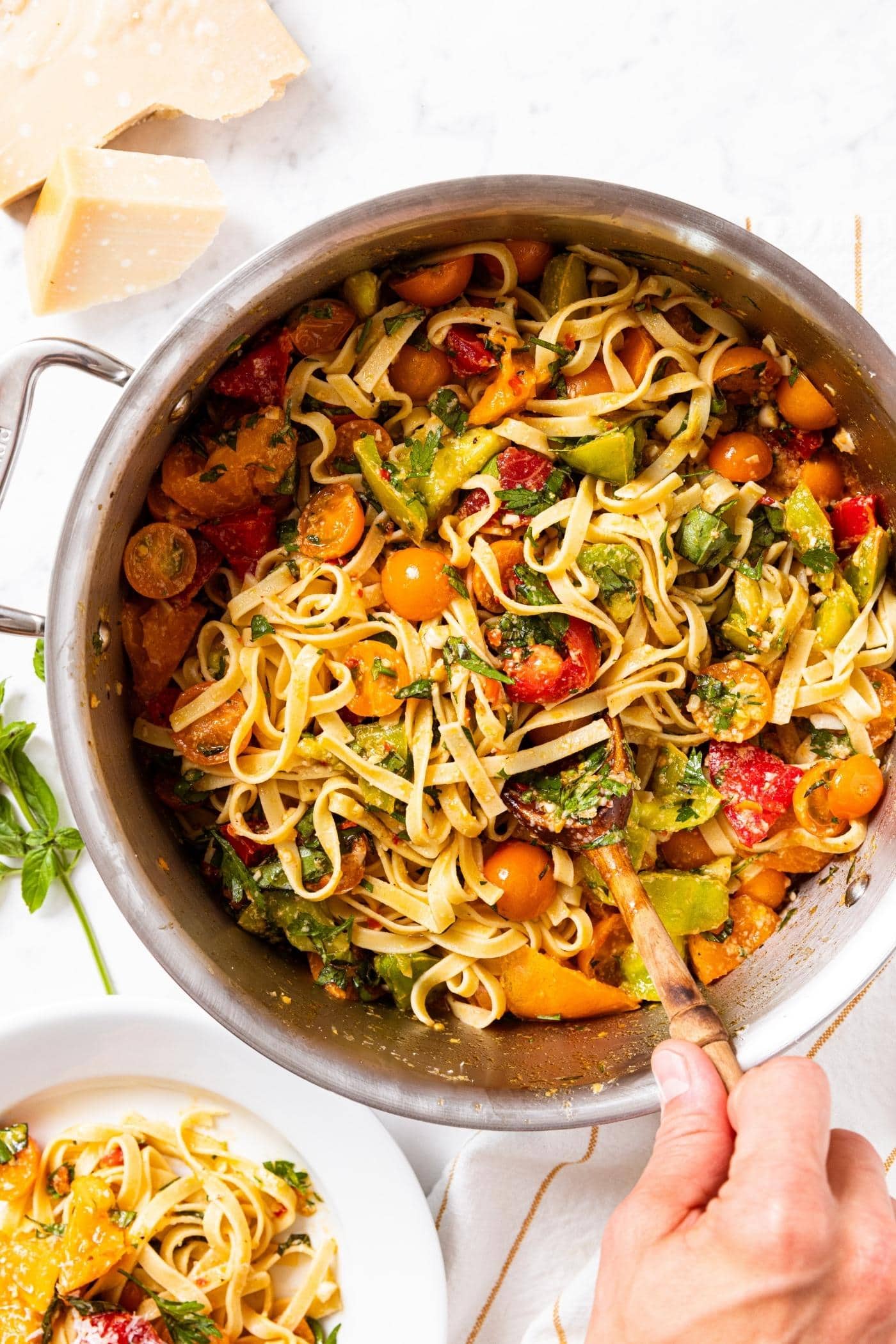 Hand stirring together pasta with tomatoes and herbs in large metal pot sitting on white countertop with Parmesan cheese to the side.