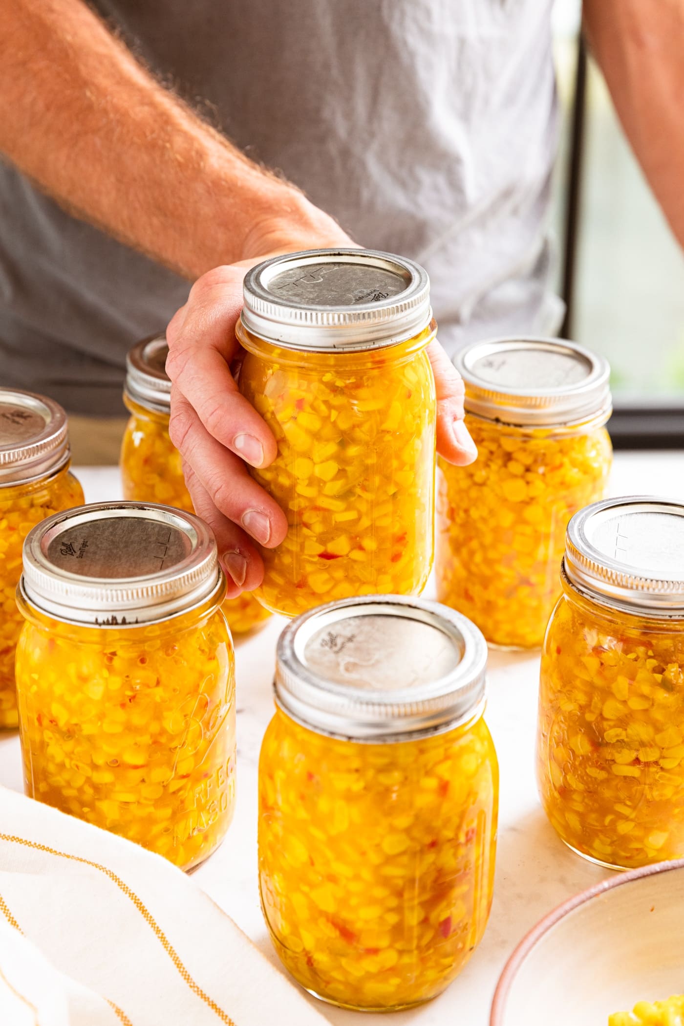 Hand holding glass jar filled with yellow colored corn relish sitting on white countertop.