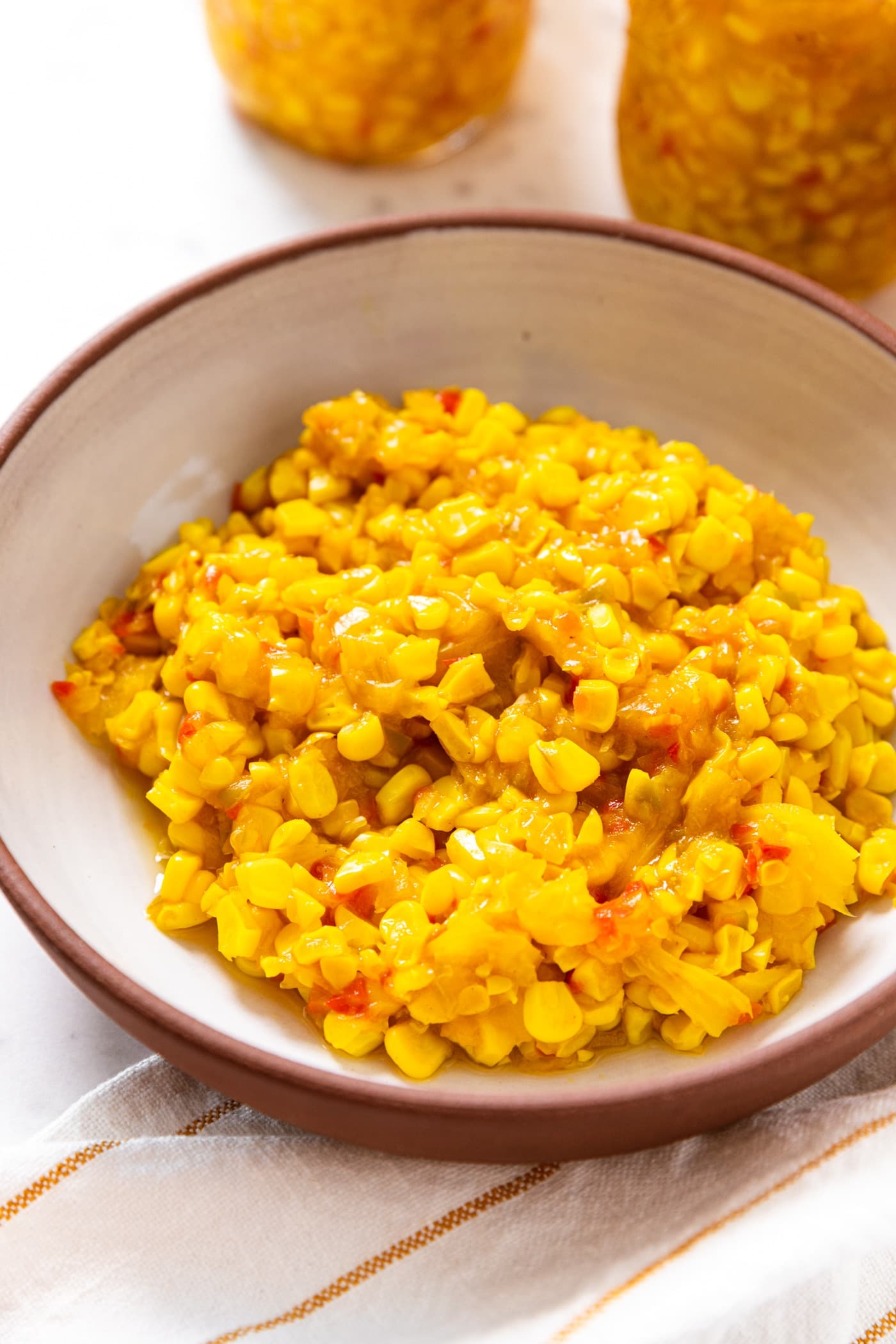 Brown bowl with white interior filled with corn relish with red bell pepper with jars in background.
