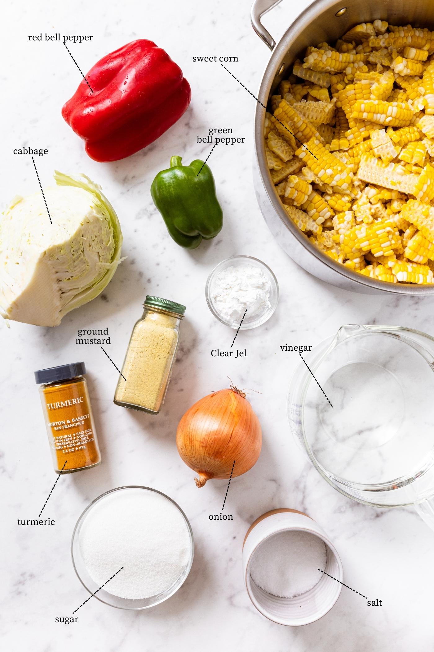Marble countertop filled with all ingredients needed to make corn relish including sweet corn, red and green bell peppers, onion, spices, cabbage, sugar, and more.