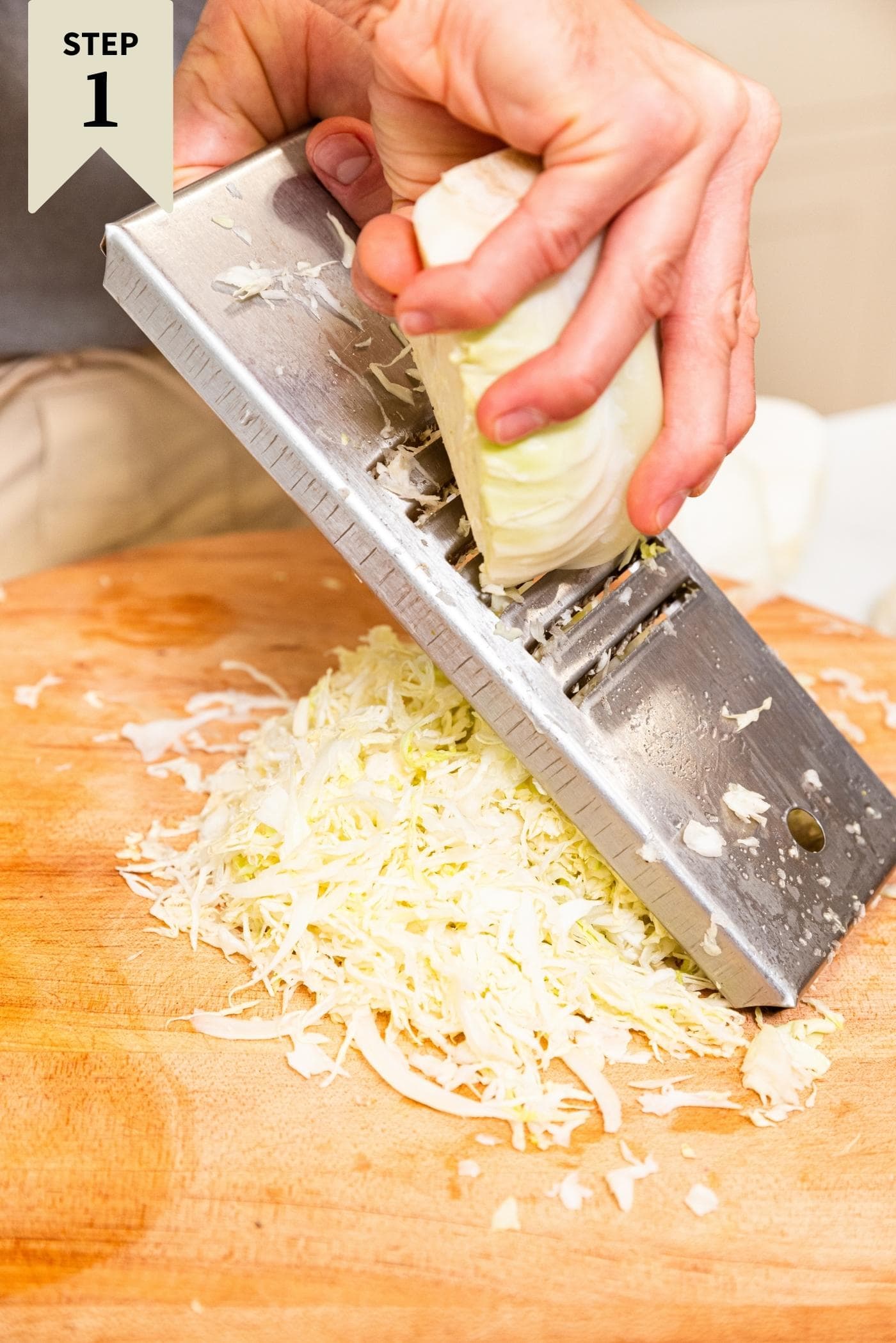Hand cutting cabbage on shredder with pile of shredded white cabbage underneath on wood cutting board.