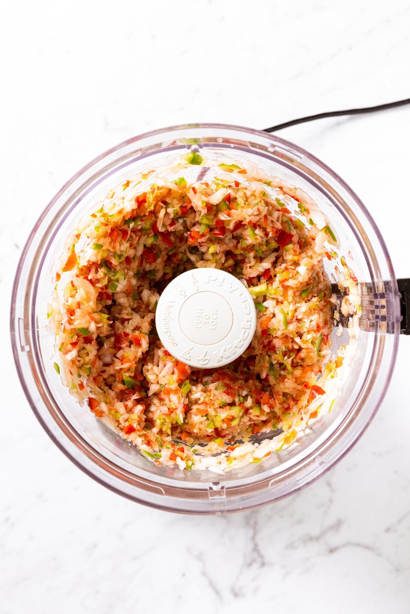 Plastic food processor with chopped up bell pepper and onion sitting on marble countertop.