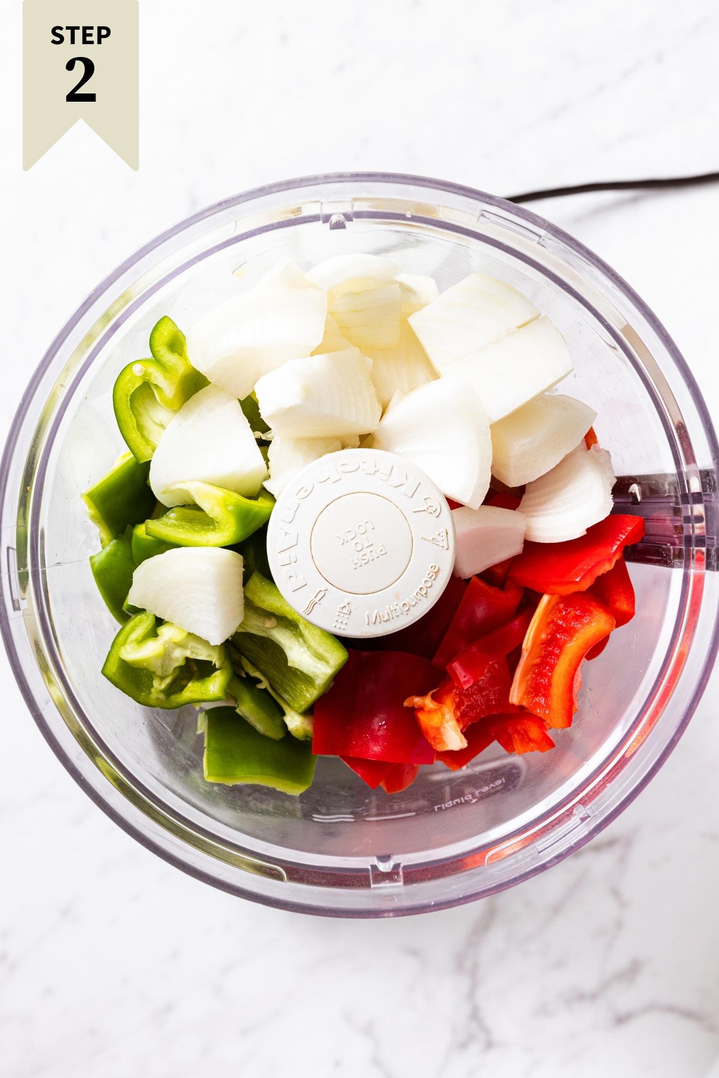 Plastic food processor bowl with green and red bell pepper and onions sitting on marble countertop in food processor.