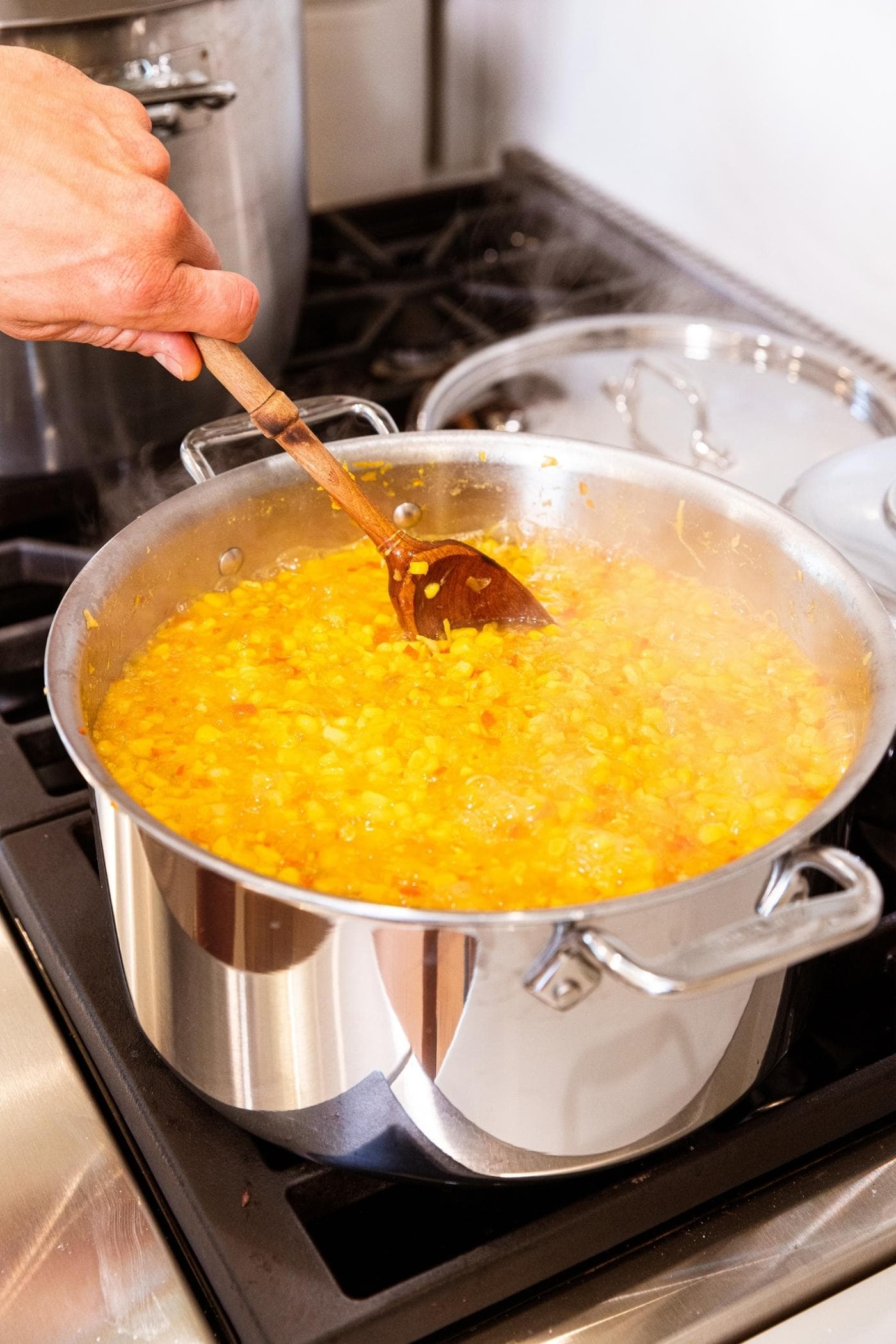Hand stirring wooden spoon in yellow corn mixture in metal kettle on stovetop.