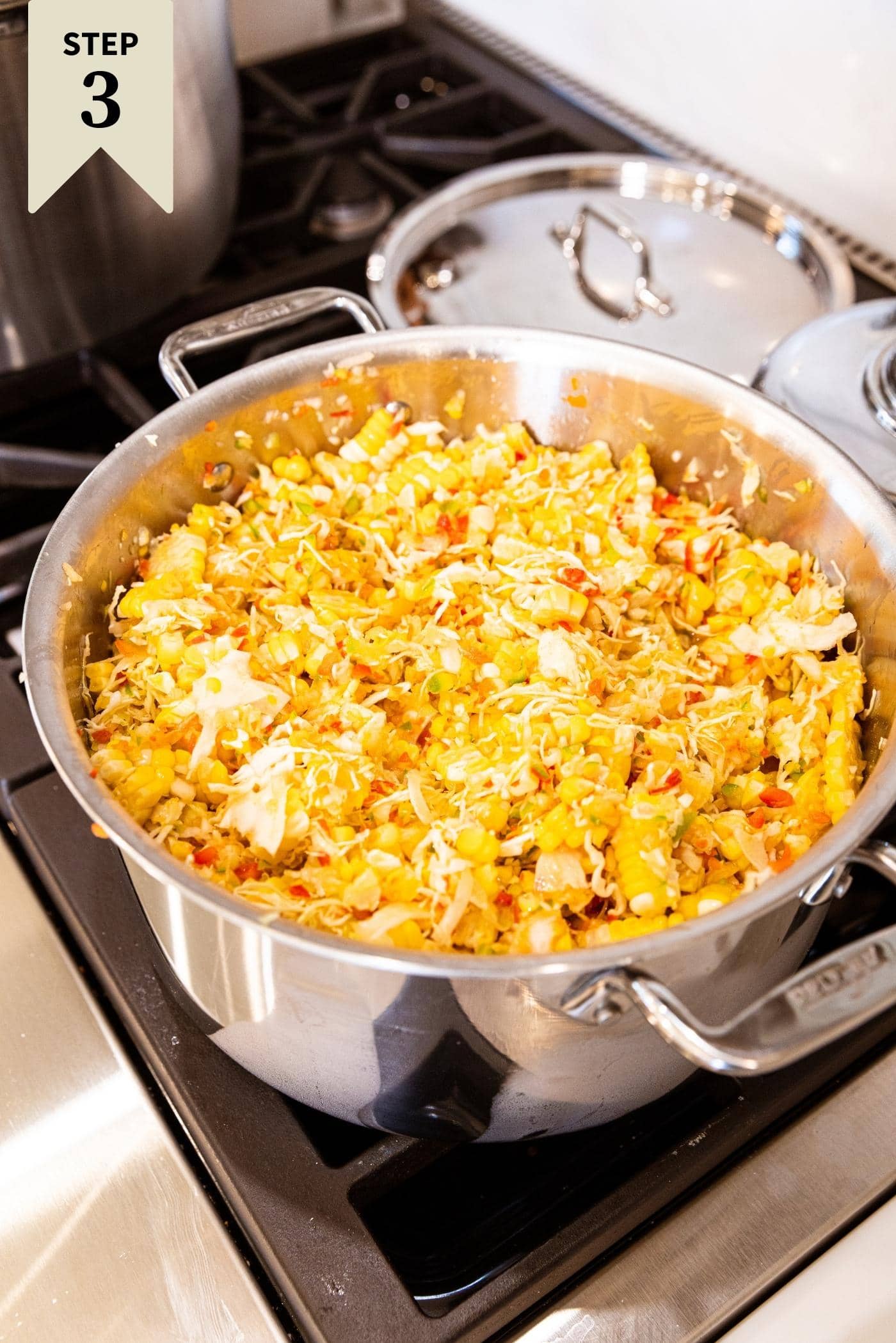 Metal stockpot sitting on stovetop filled with corn, cabbage, onion, and bell pepper.