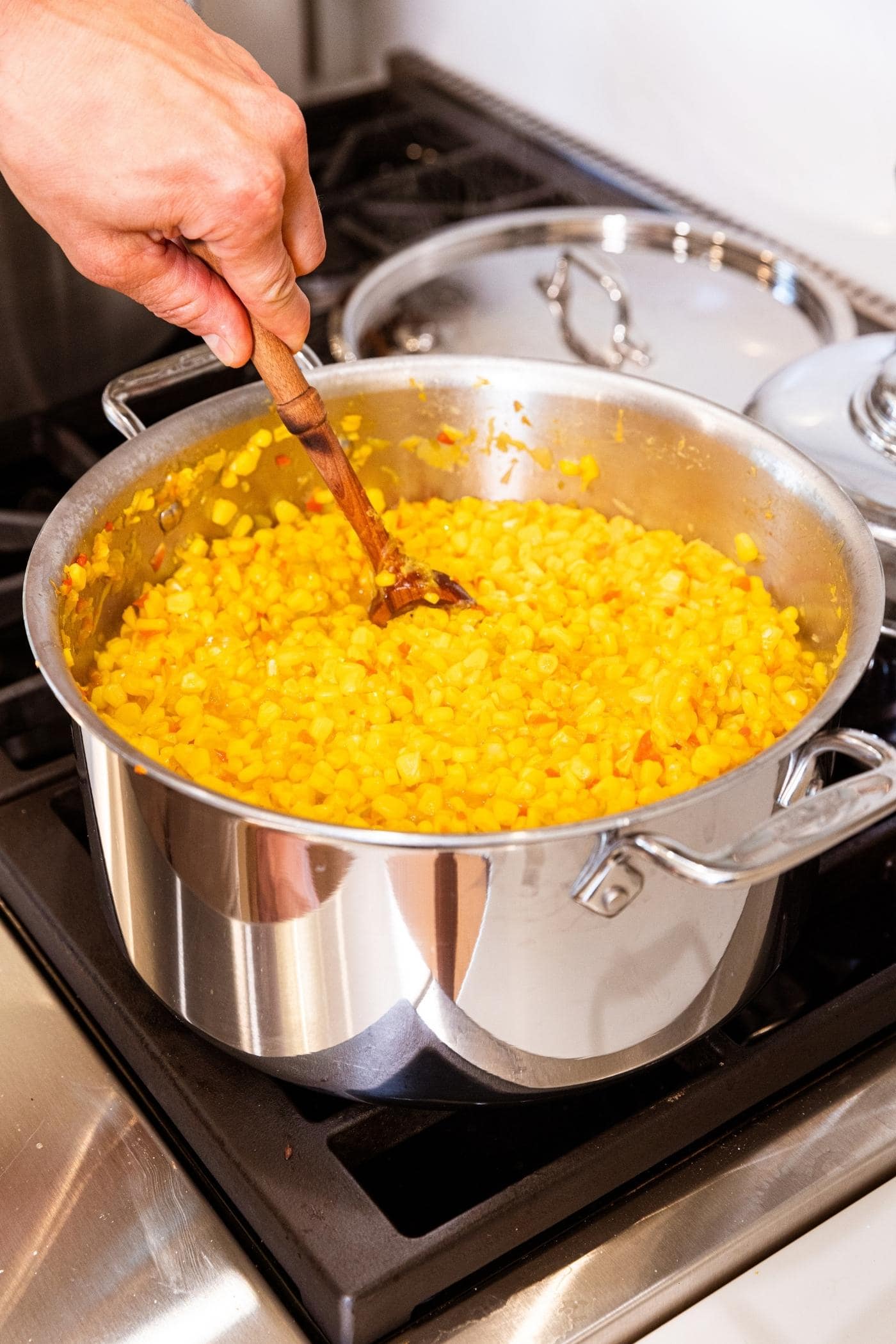 Hand stirring corn relish mixture with wooden spoon in large pot on the stovetop with lid of pot in the background.