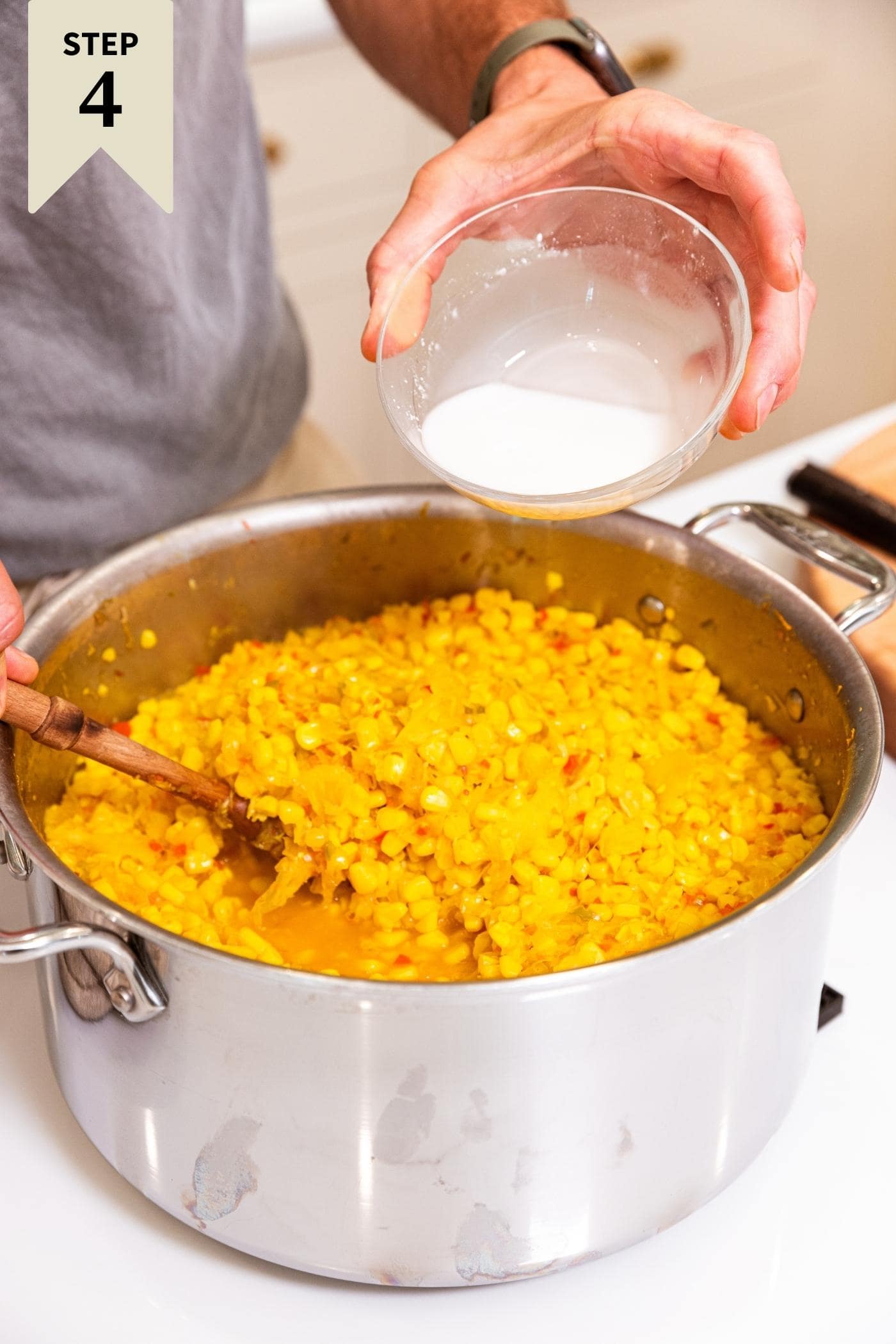 Hand pouring white slurry into metal stockpot filled with corn relish sitting on white countertop.
