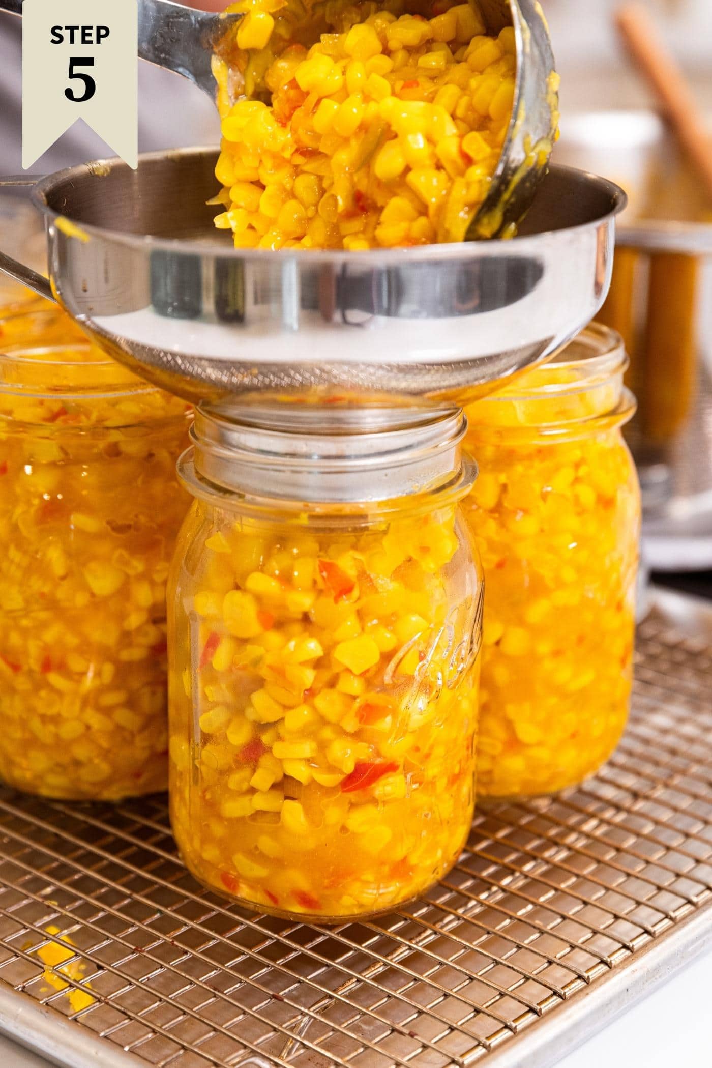 Hand ladling corn relish into glass jar with funnel attachment.