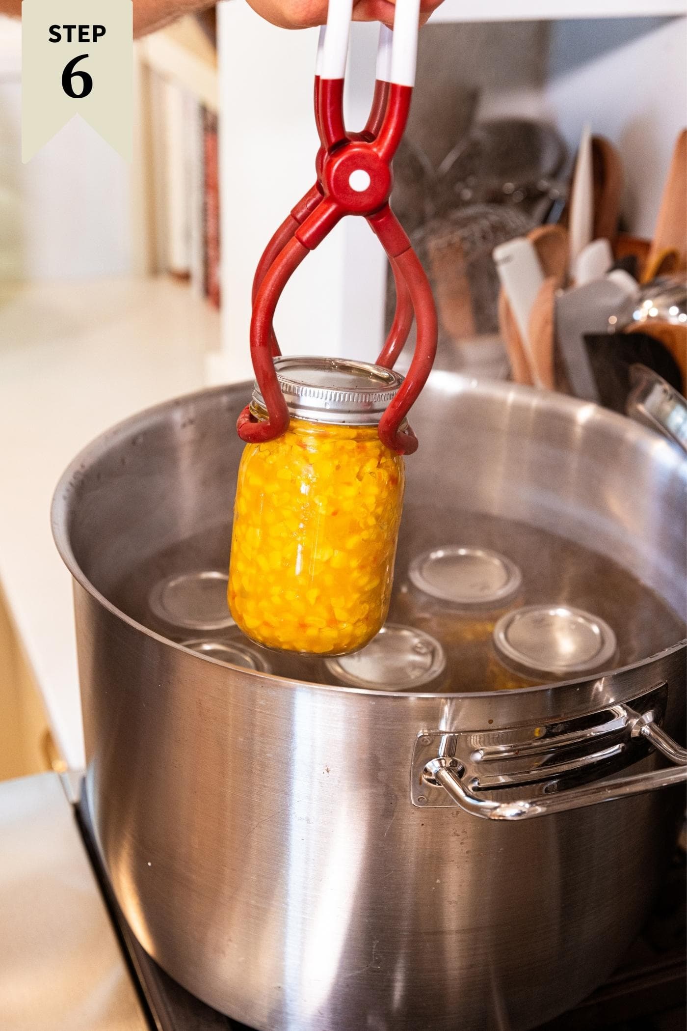 Jar lifter putting glass jar filled with corn relish going into metal stockpot on stove.
