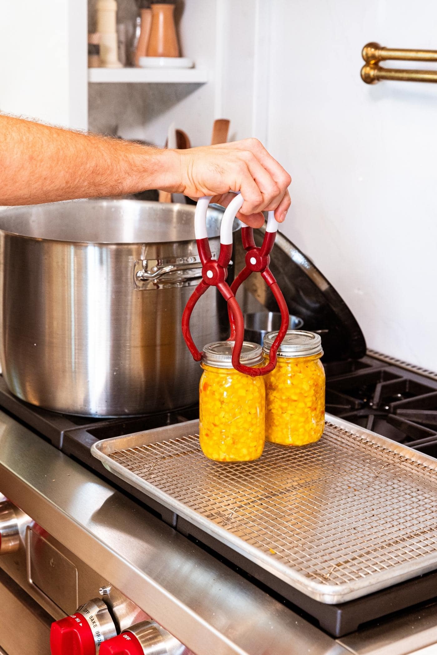 Hand holding jar lifter putting glass jar filled with corn relish removing from stockpot onto a wire rack.