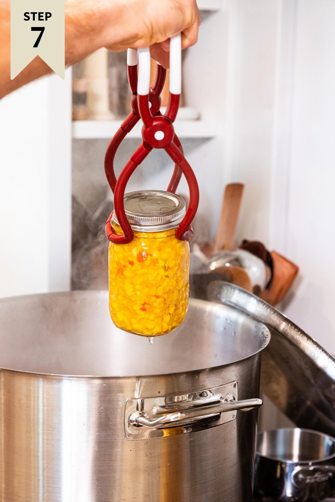 Hand holding jar lifter holding glass jar filled with corn relish pulling from metal stockpot on stove.