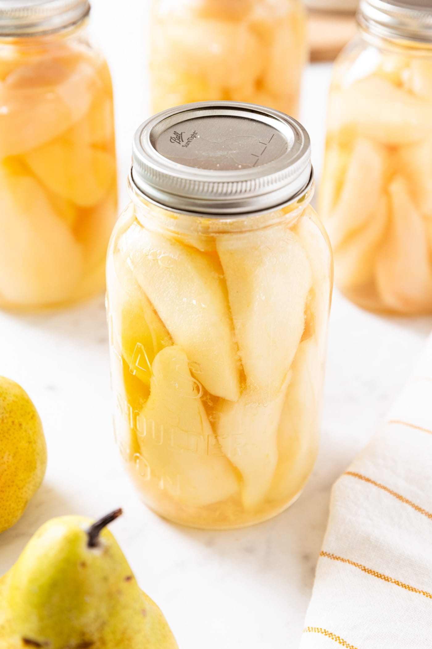 Glass jars filled with sliced canned pars sitting on marble surface with more unsliced pears as well.
