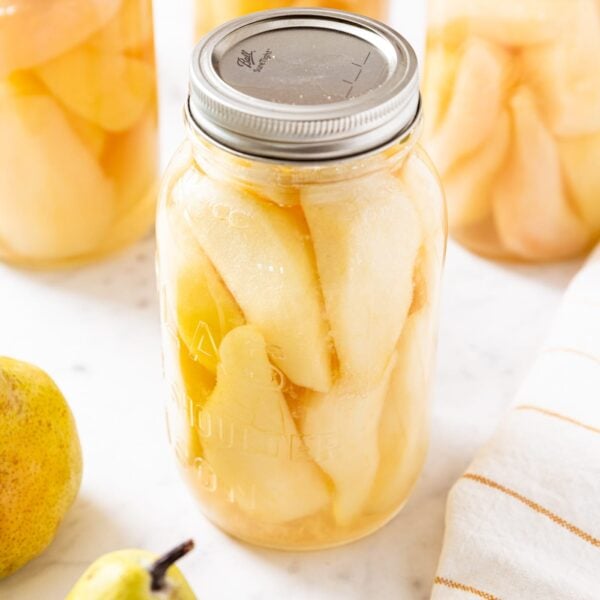 Canned jar of pears sitting on marble surface with more jars in background as well as some unsliced pears.