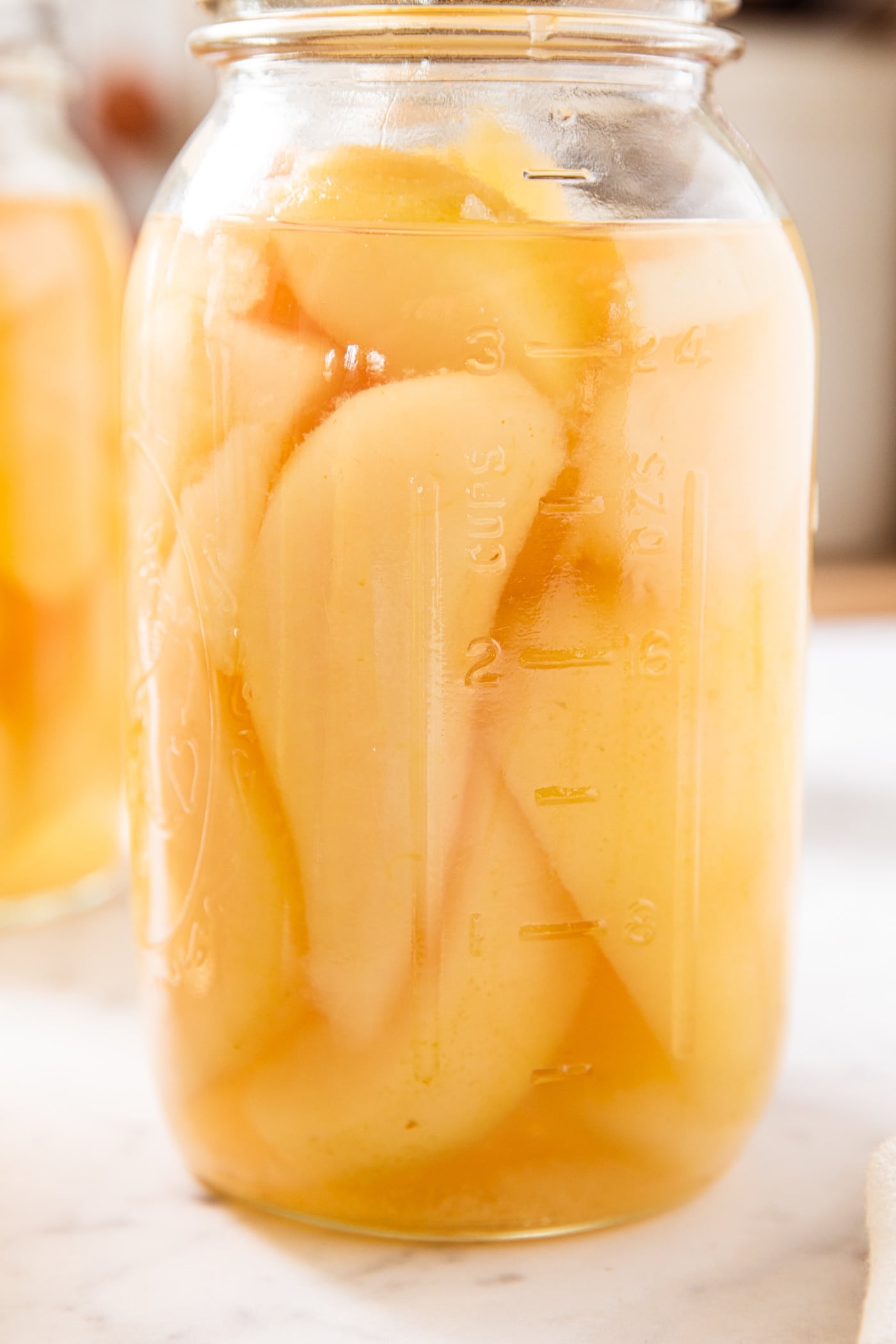 Glass jar filled with slices of canned pears with liquid surrounding the pears sitting on white marble surface with more jars in background.