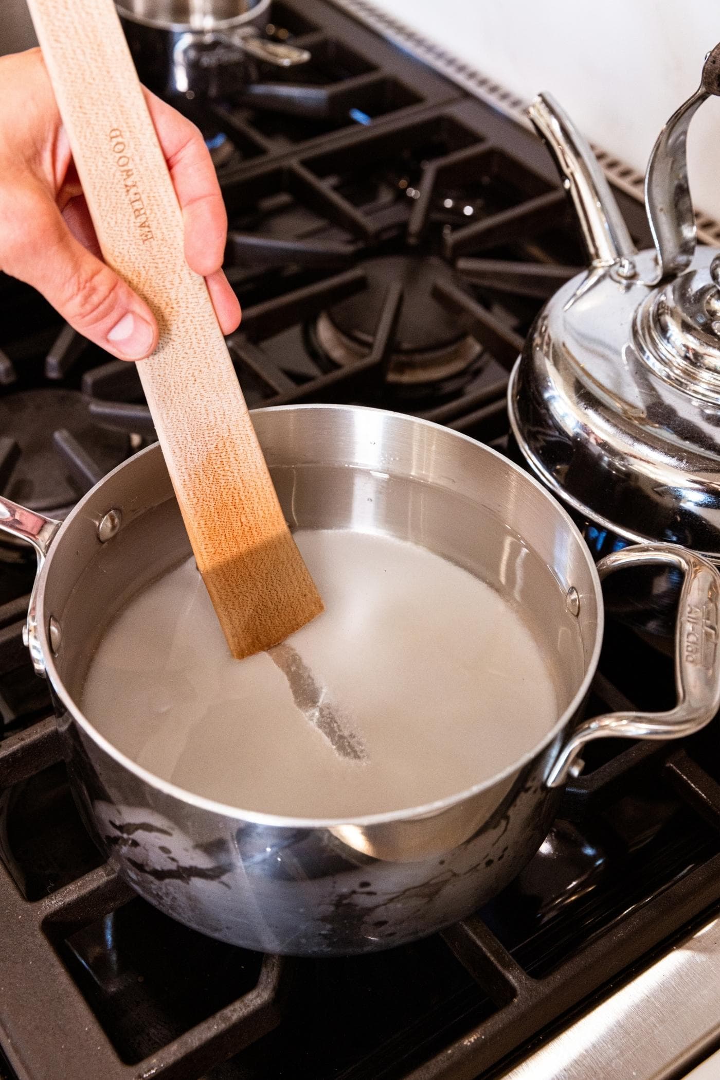 Metal kettle sitting on stovetop with sugar in the bottom with water on top and wooden spatula scraping the bottom.