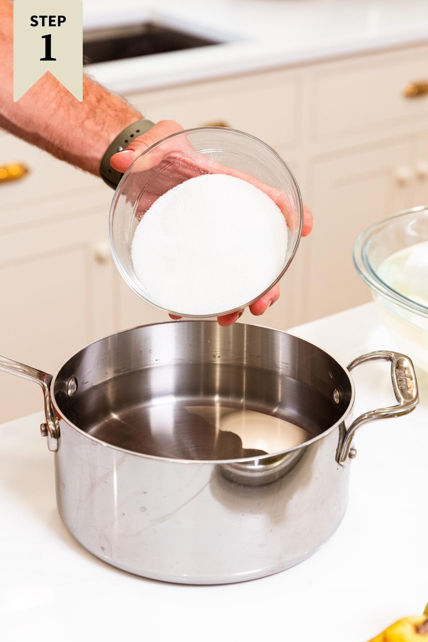 Hand pouring sugar into a metal kettle filled with water from glass bowl with white countertop and cream cabinets in background.
