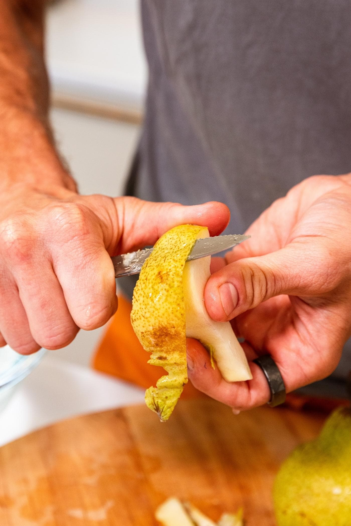 Two hands holding sliced pear cutting off the skin with paring knife over cutting board with more pears below.