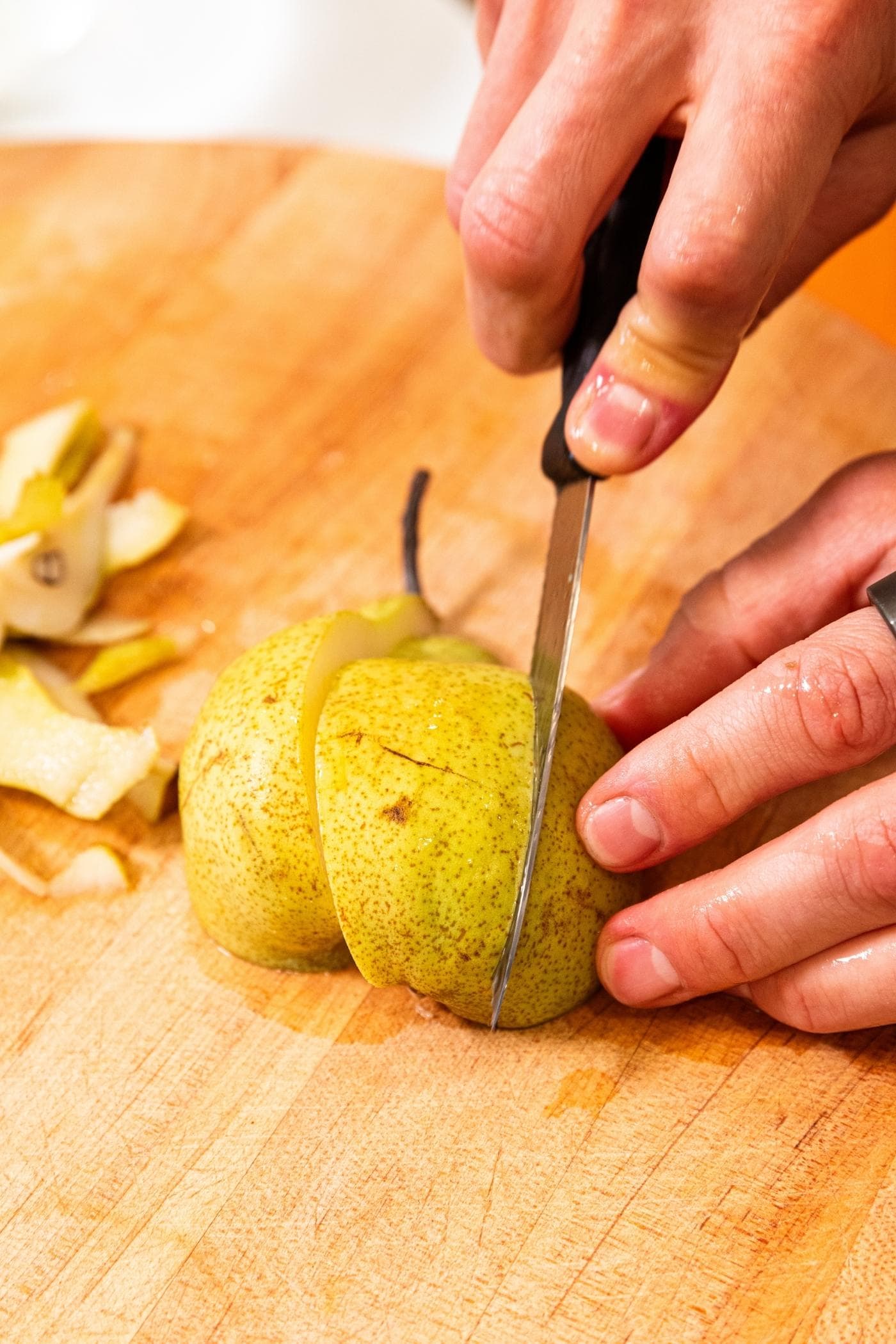 Hand slicing half of pear into thirds still with skin on all on wooden cutting board.