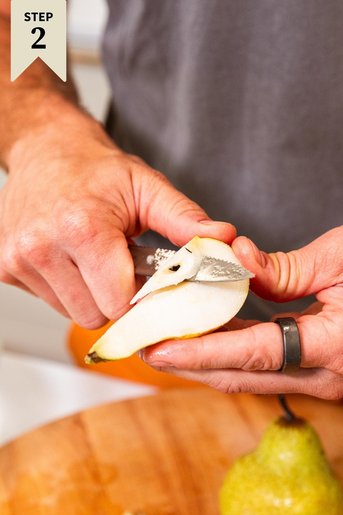 Hand slicing core from sliced pear in hand with wooden cutting board in background as well as additional pear.