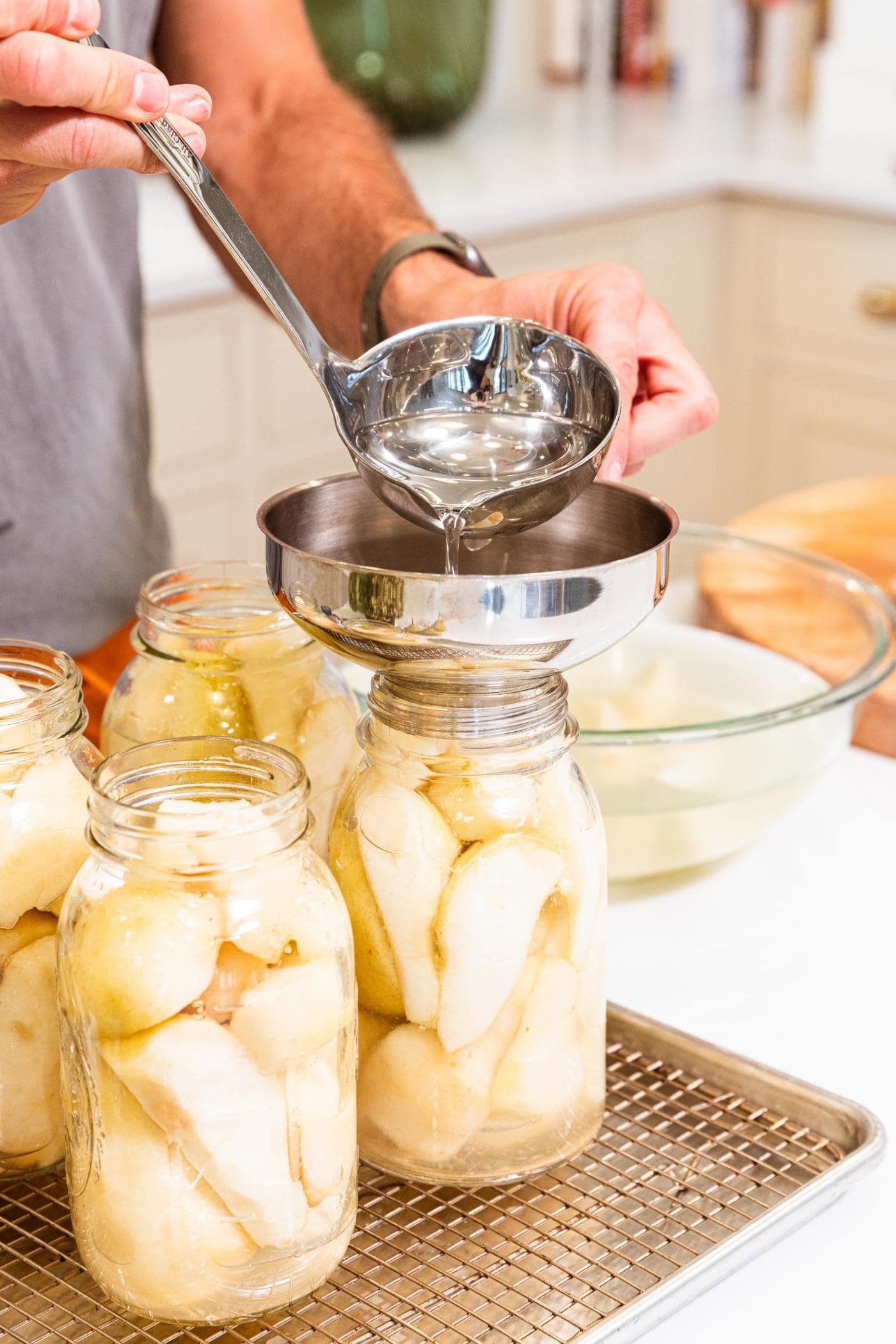 Hand using metal ladle pouring sugar syrup into glass canning jars filled with slices of pears.