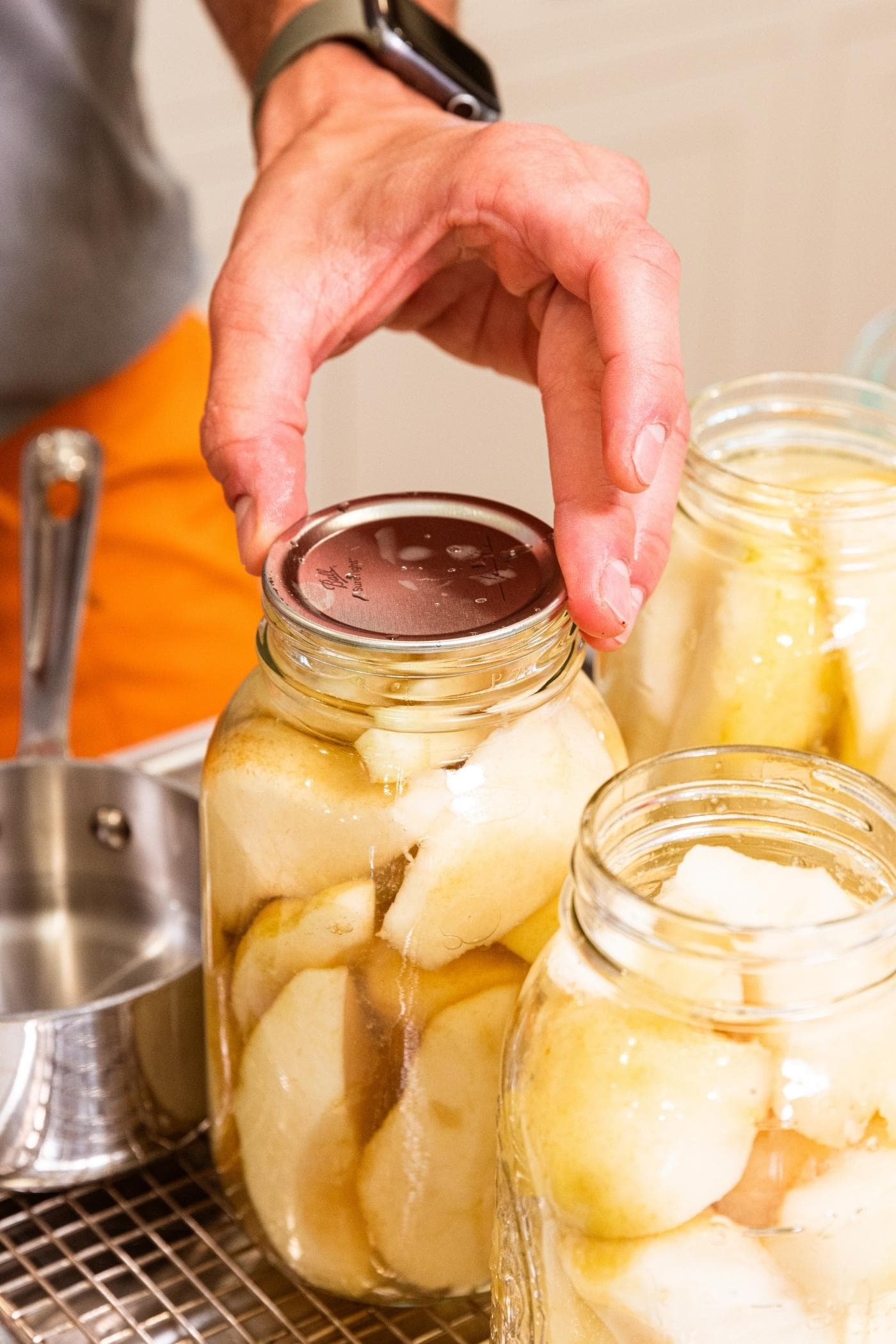 Hand placing lid on top of canning jar filled with slices of pears on metal cooling rack.