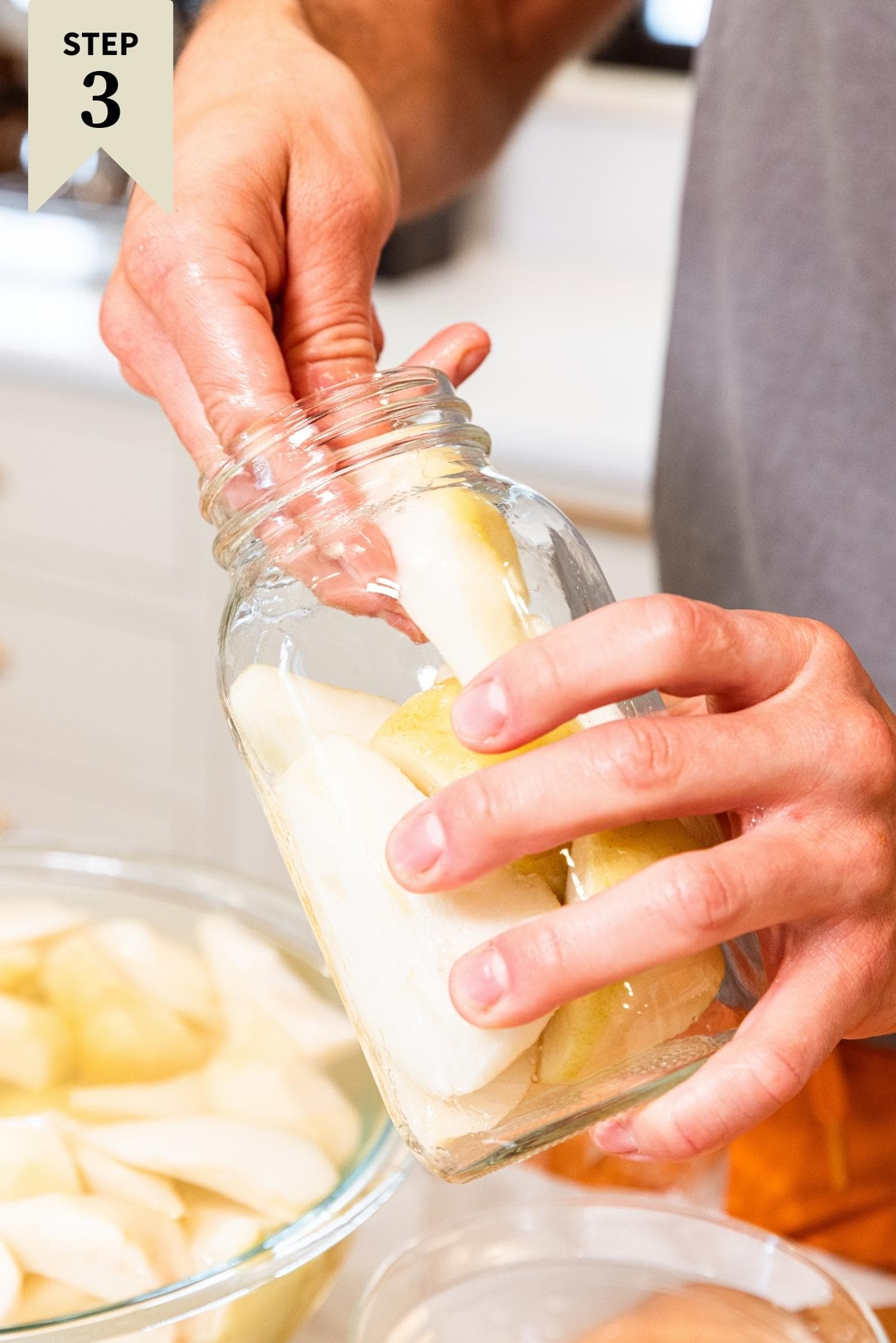 Hand holding glass canning jar fitting slices of pears into jar with more pears in bowl of water in background.