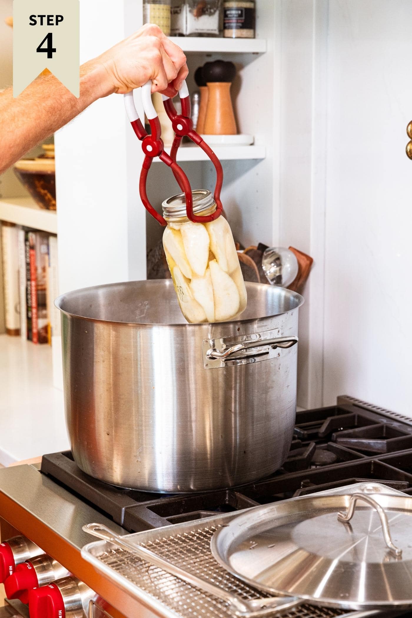 Hand placing glass canning jar in metal water bath on stovetop.