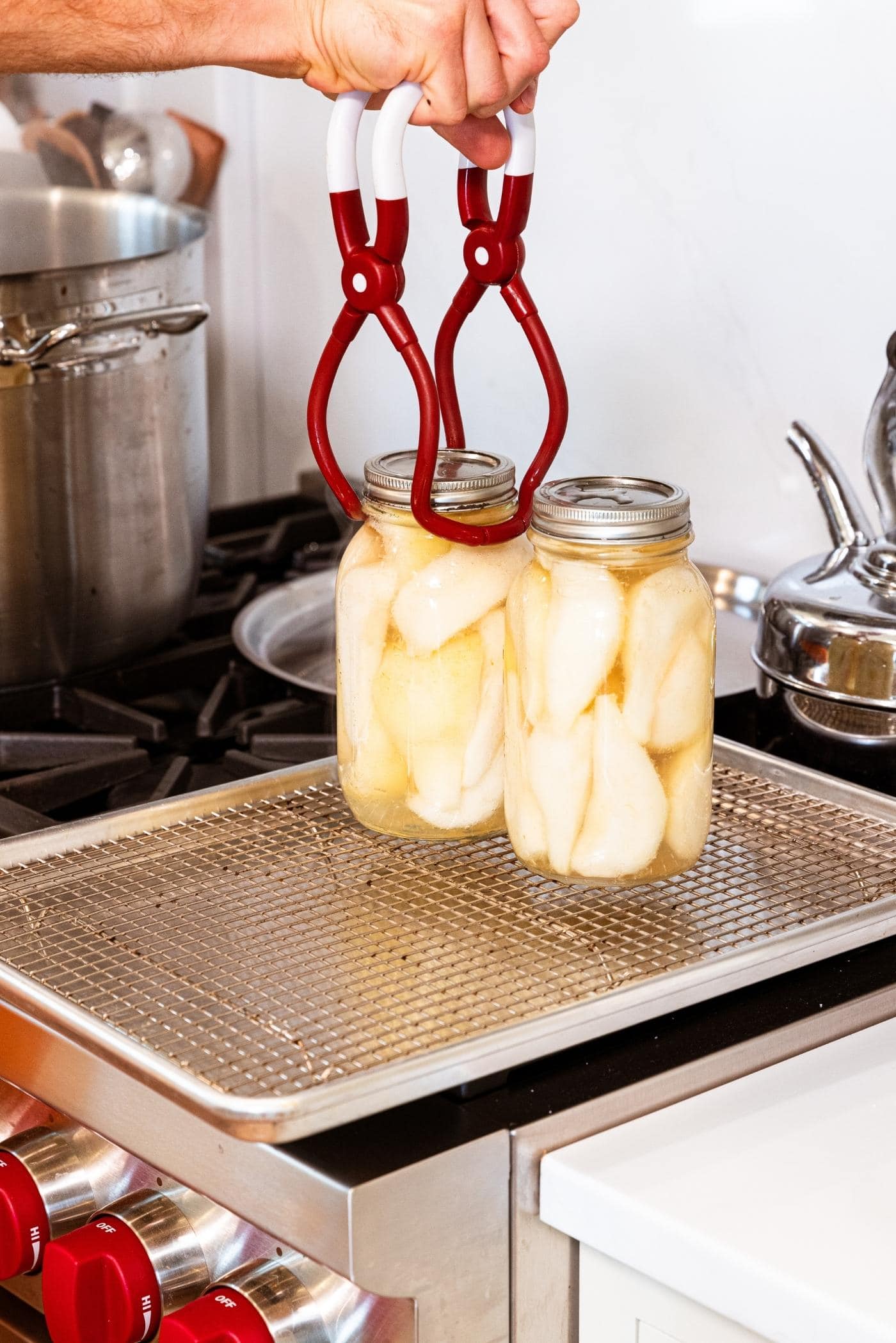 Hand placing glass canning jars filled with pears on metal rack on stove with clamps.